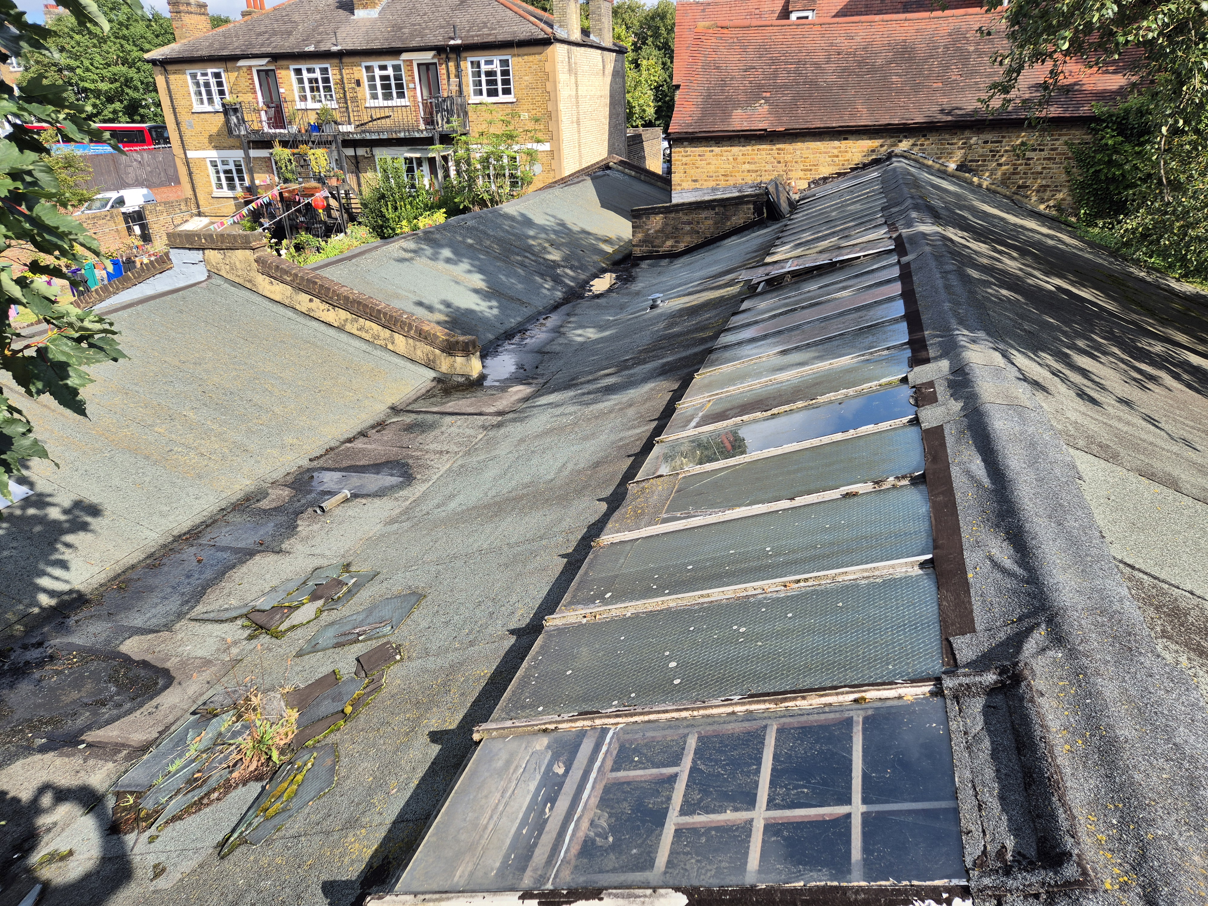 Flat roof under renovation with exposed beams, glass panels, and building materials. Yellow brick houses visible in background.