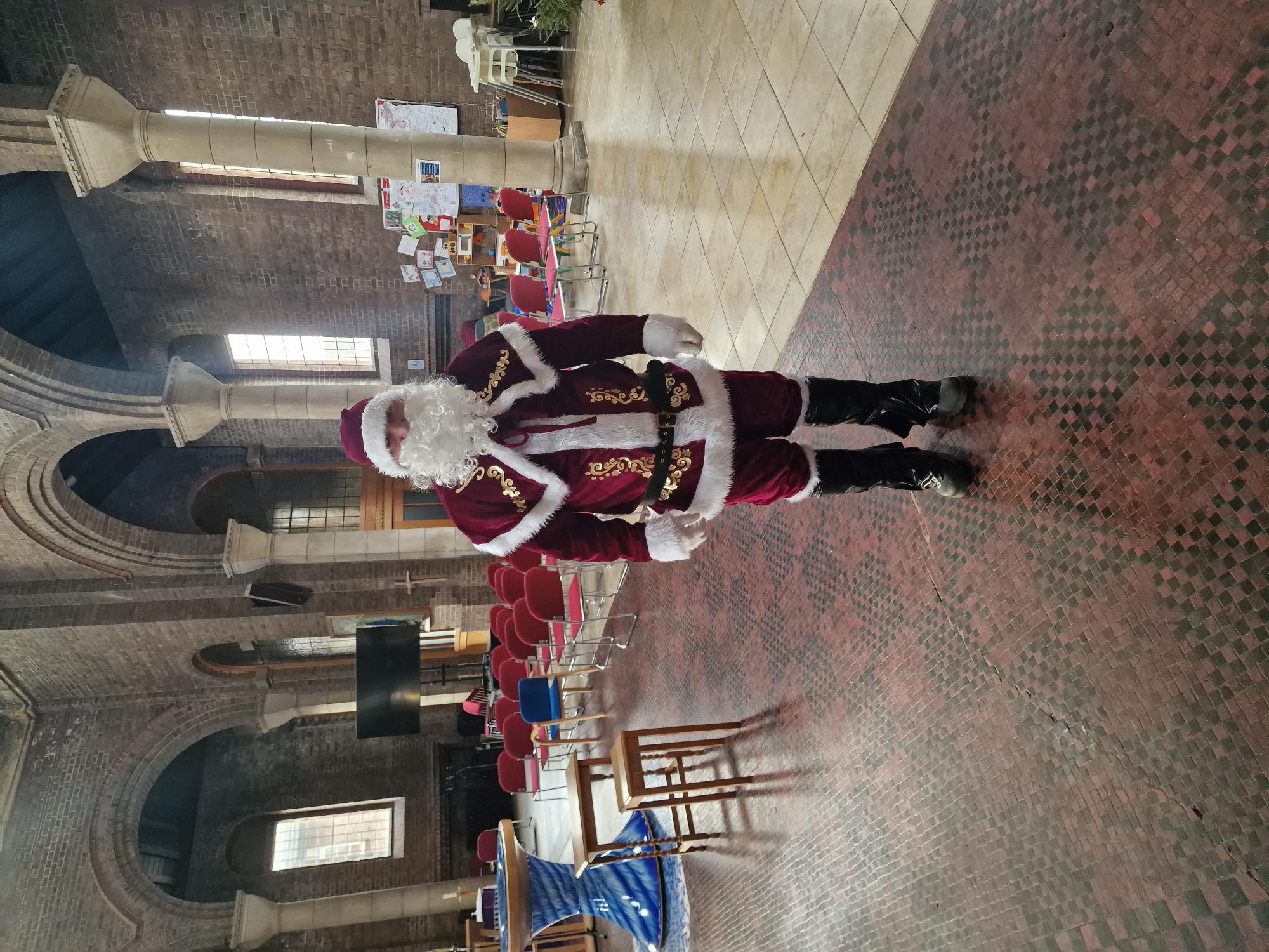 Person in red and white Santa costume standing in shopping centre with tiled floor, shops and seating areas visible in background.