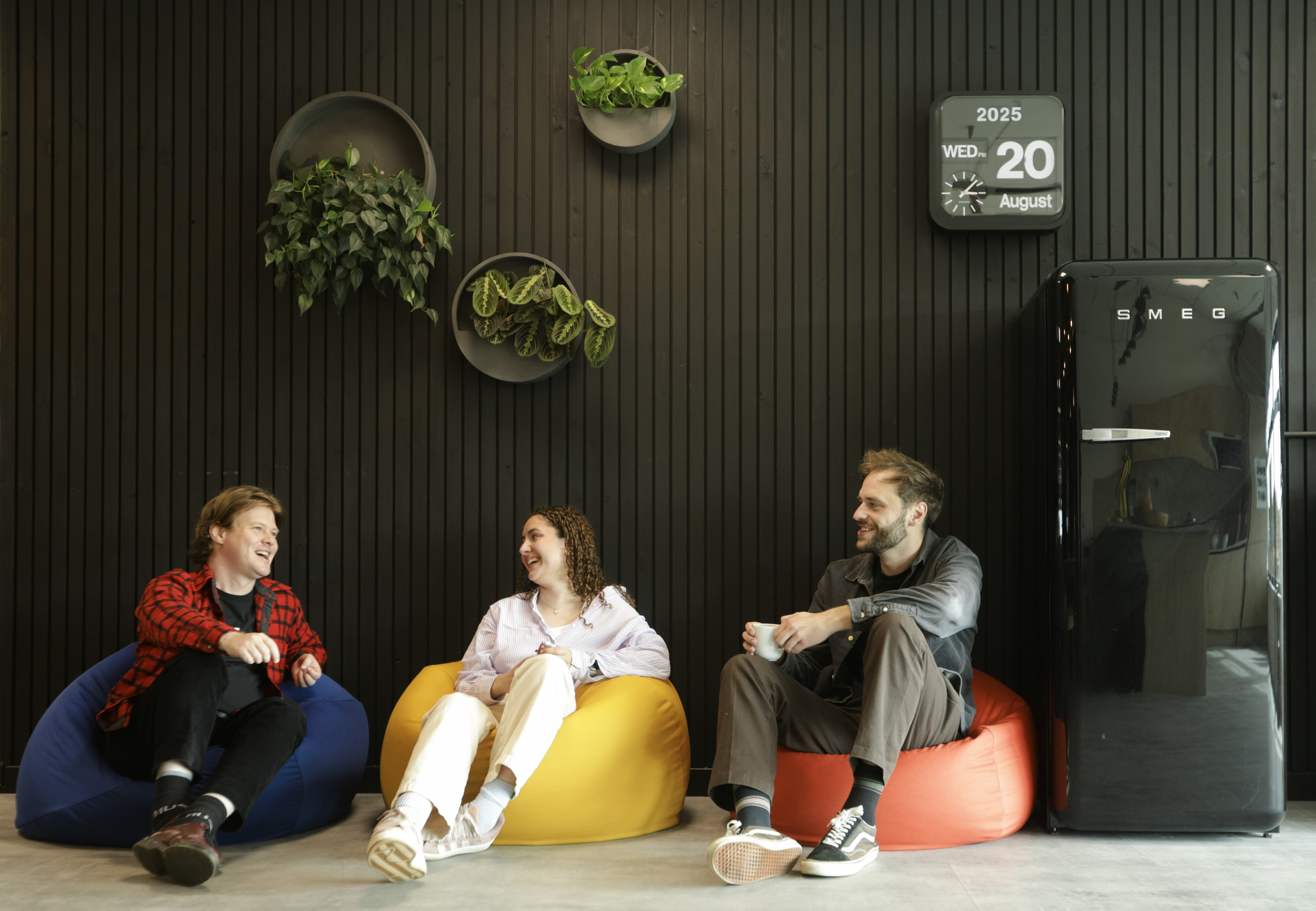 Three people sitting on colourful bean bags (blue, yellow, orange) against dark wooden wall with hanging plants and vintage fridge.