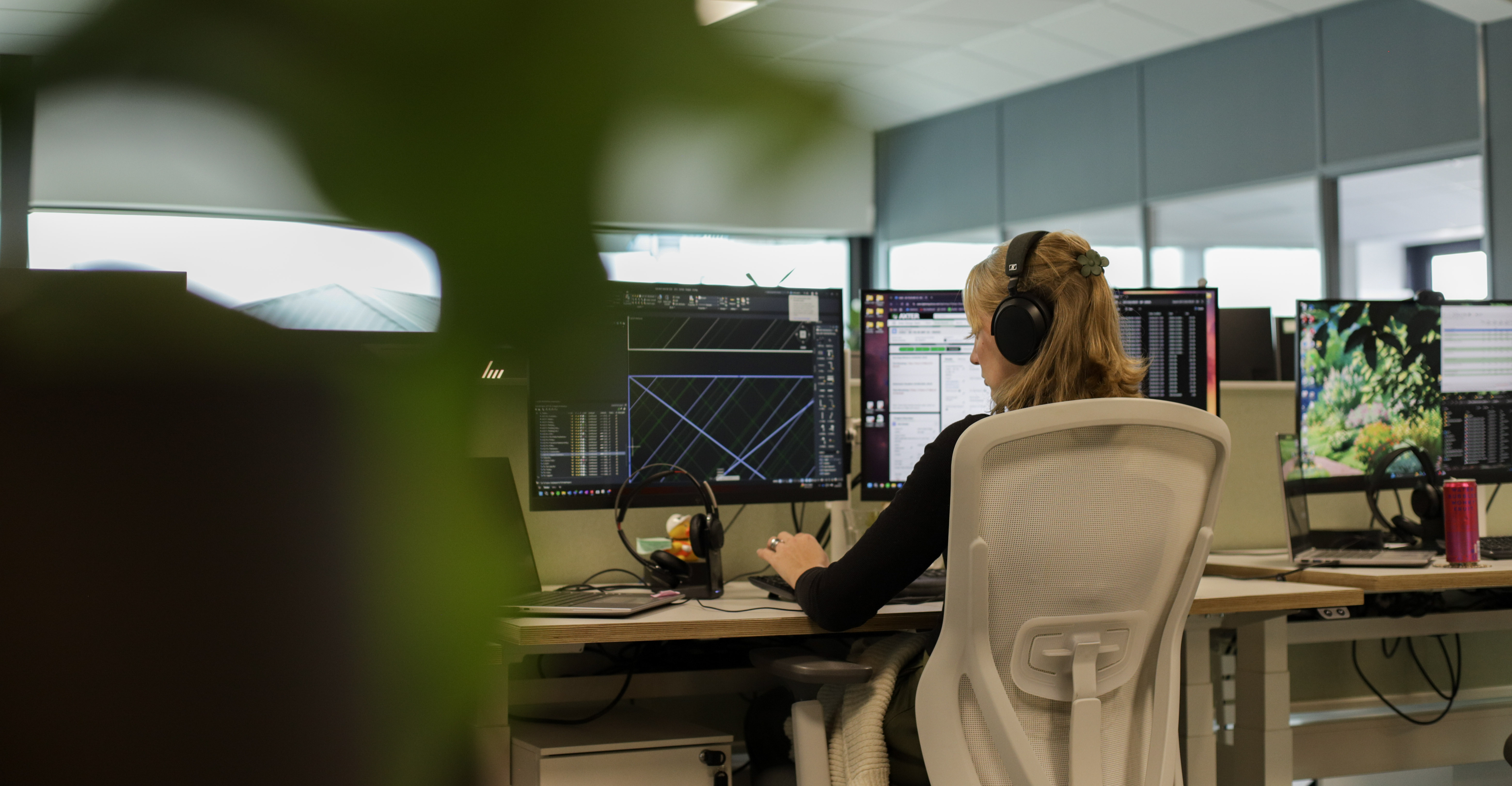 Woman with blonde hair in bun sits at wooden desk with multiple computer monitors in modern office with blue-grey walls and windows.