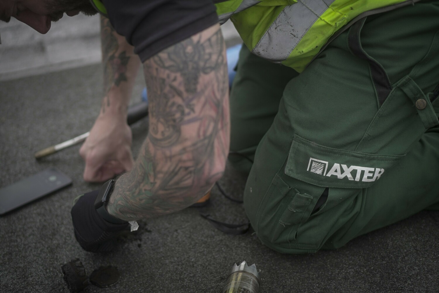 Person kneeling on pavement wearing camouflage trousers and green knee pad with "BAXTER" text, holding dark object.