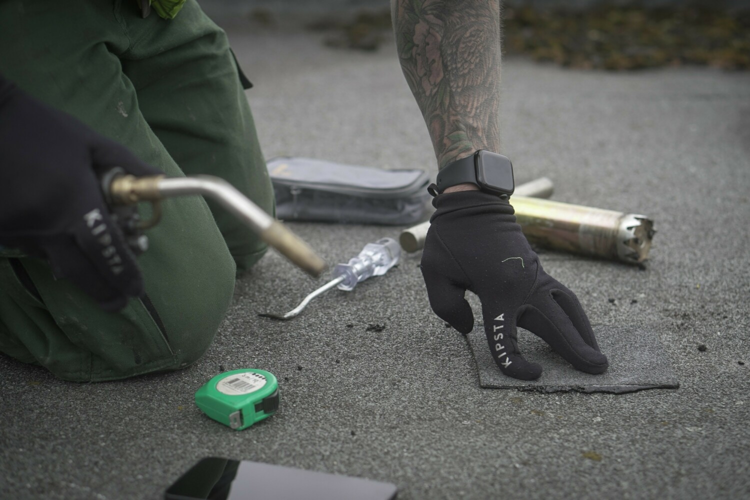 Person in green jacket and black gloves crouched on pavement, holding metal tool near green tape measure.