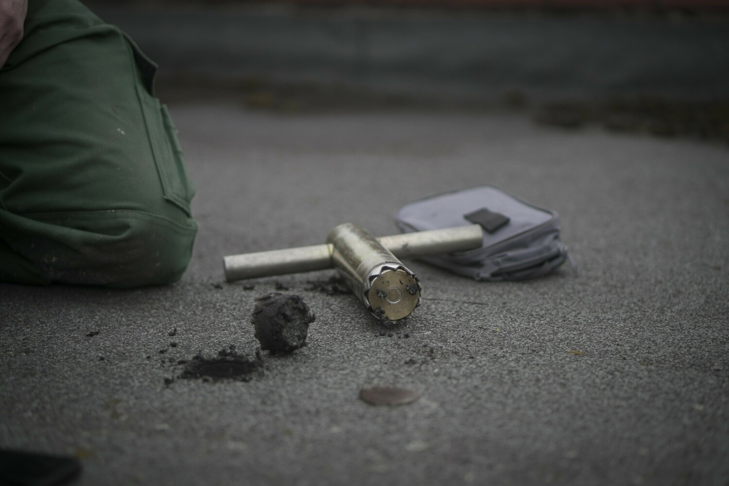 Person in green jacket sits on pavement near scattered papers and metal cylindrical object on grey asphalt surface.