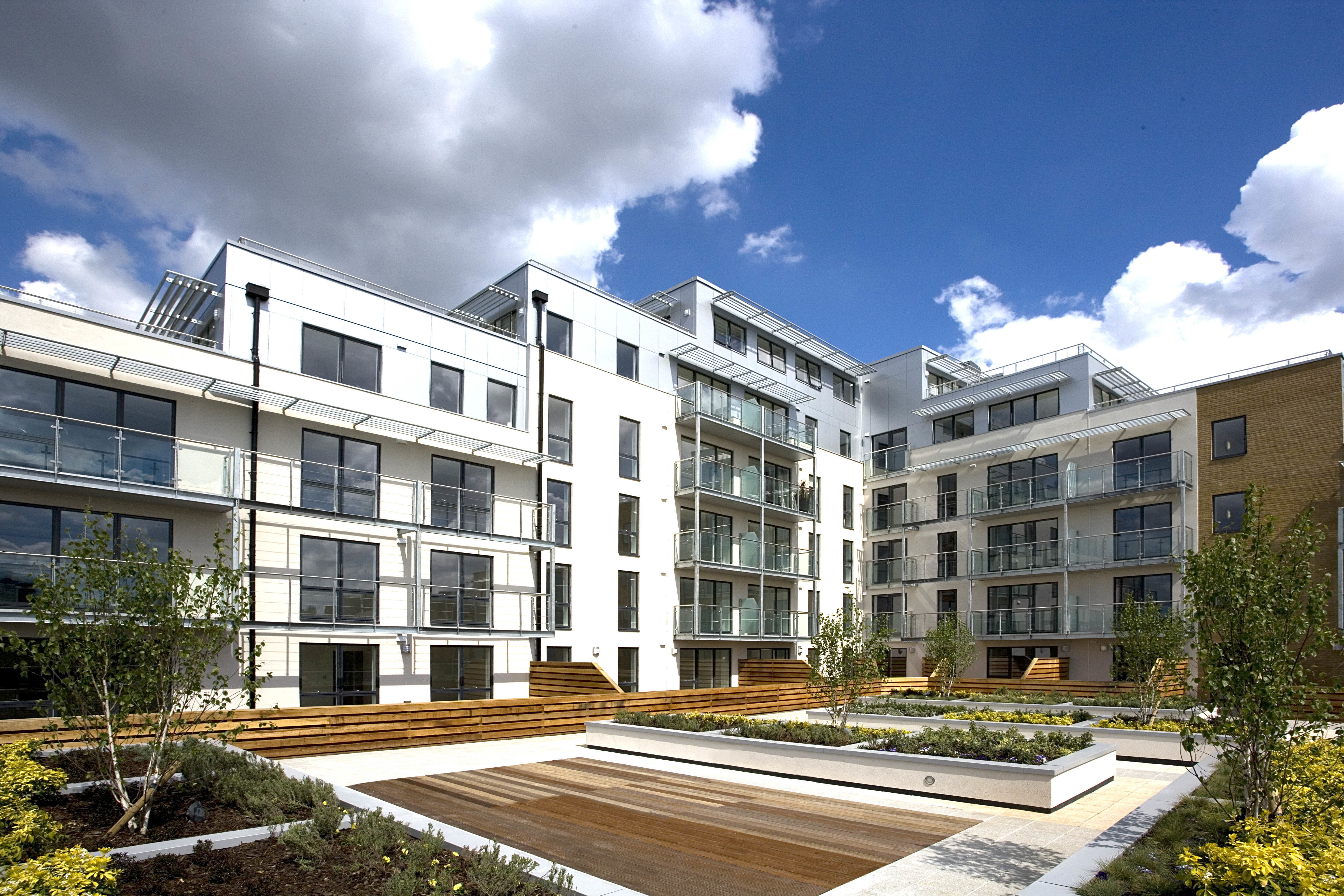 Modern white residential buildings surrounding landscaped courtyard with wooden decking, planted areas, and glass balconies under blue sky.