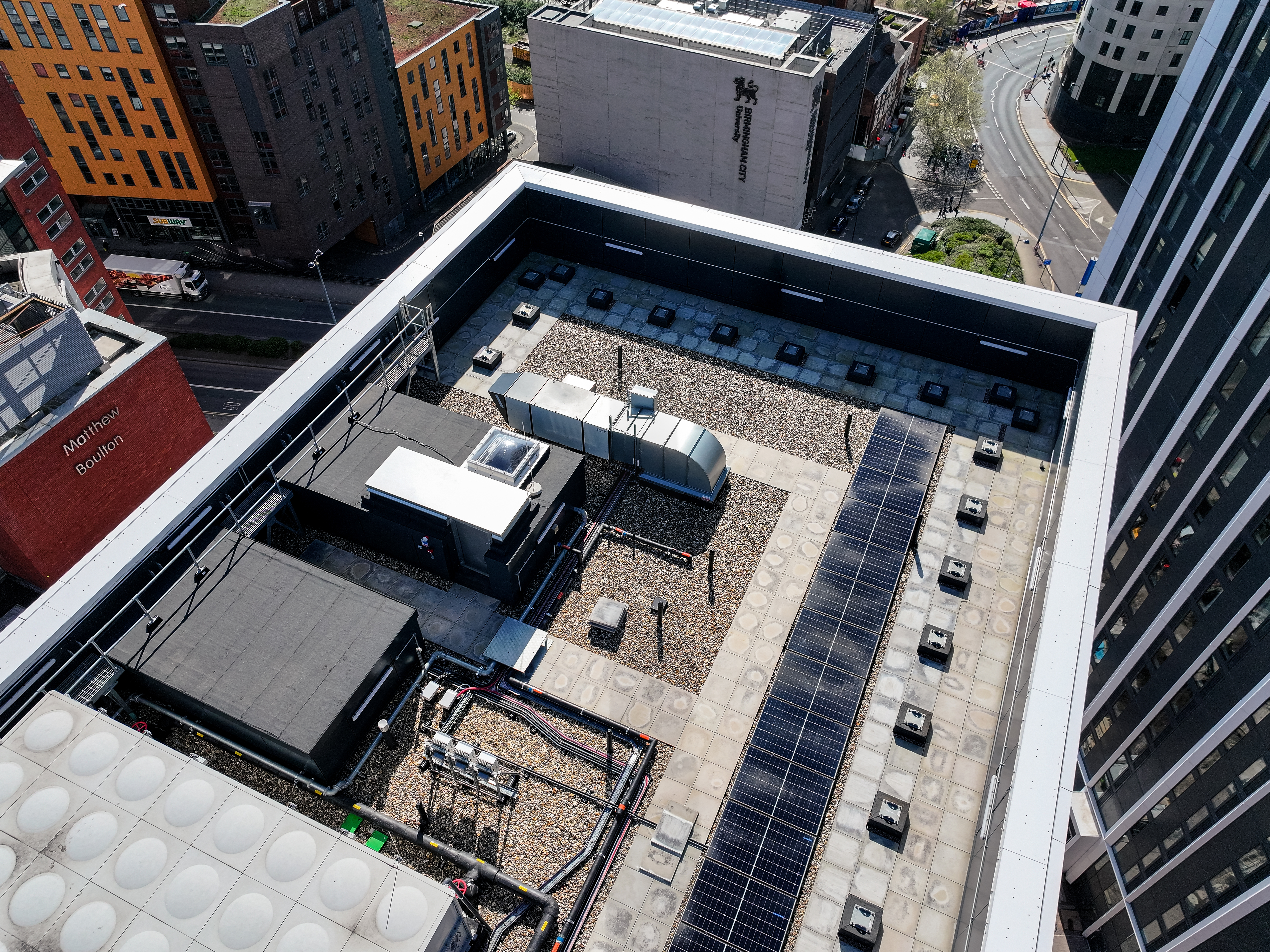 Aerial view of a flat urban rooftop with solar panels, HVAC units, and mechanical equipment, surrounded by city buildings and a road.