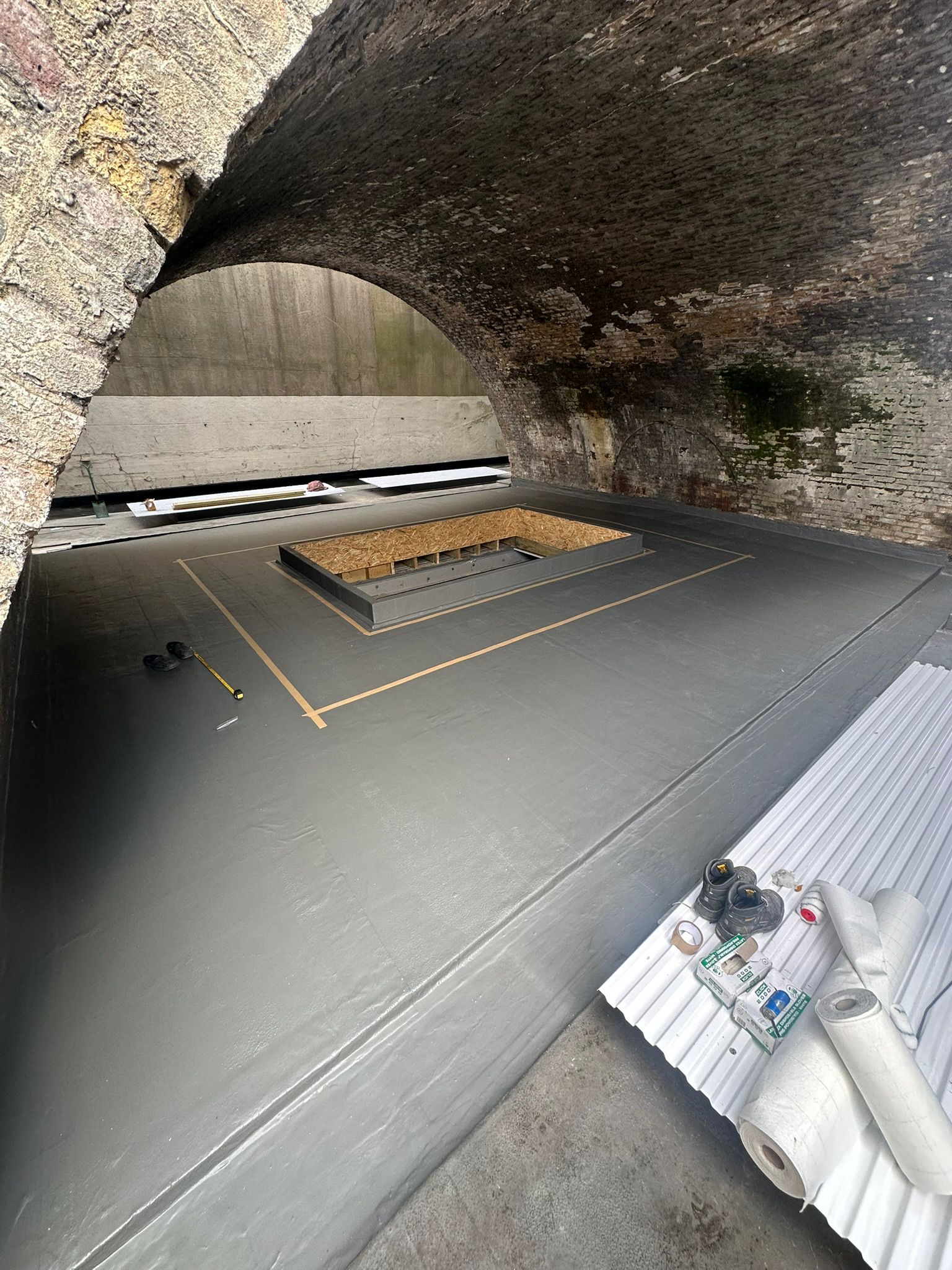 Stone arched tunnel interior with grey floor, wooden platform structure in centre, white equipment and papers on right side.