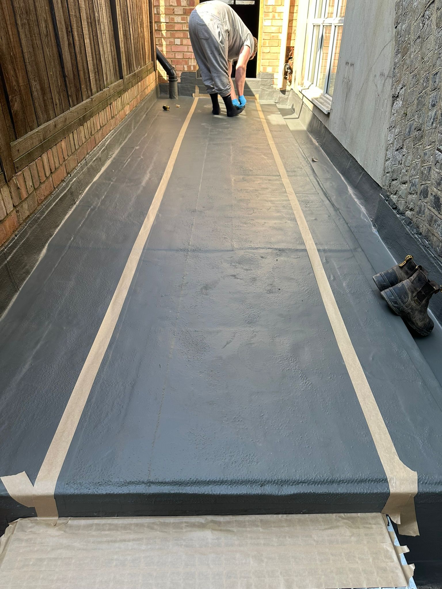 Man laying wooden strips on grey concrete floor between brick walls, creating parallel lines for flooring installation.
