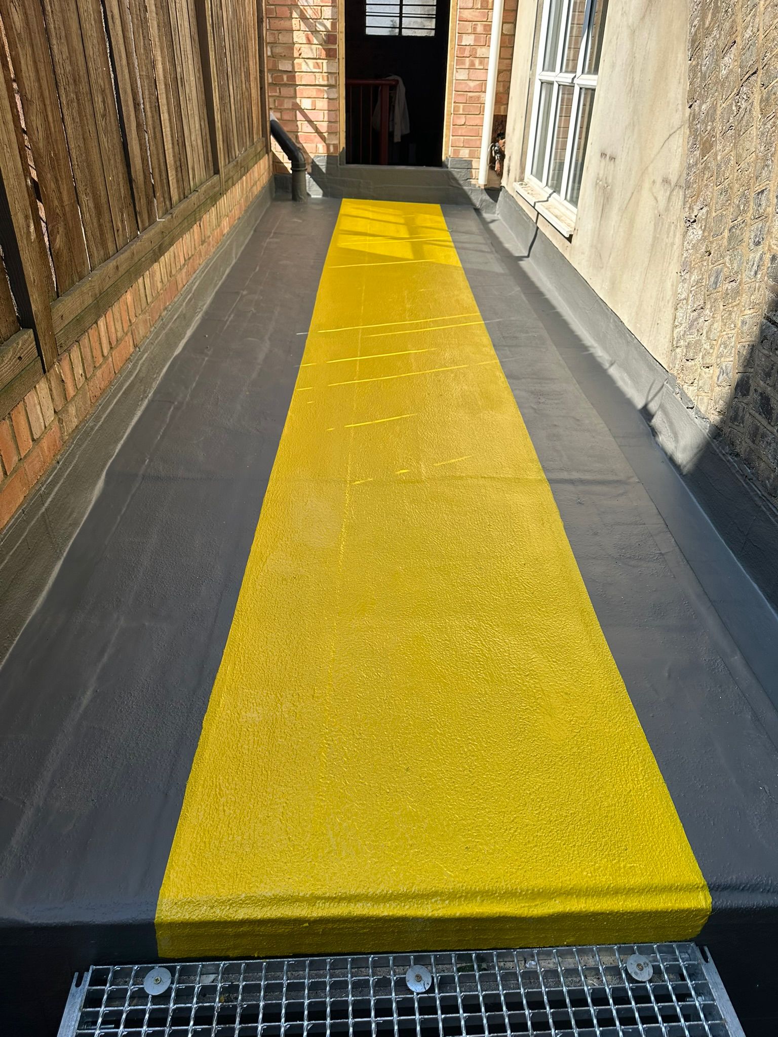 Bright yellow tactile paving strip runs down centre of grey concrete ramp between brick buildings, with metal grating at bottom.