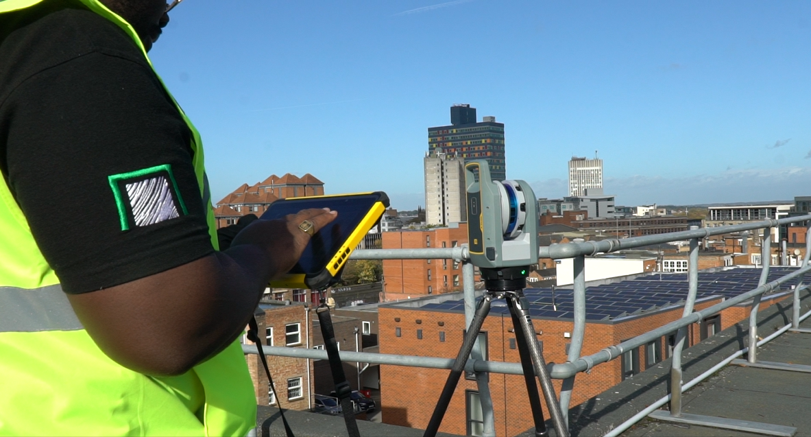 Surveyor in high-vis jacket using surveying equipment on tripod overlooking urban area with brick buildings and clear blue sky.