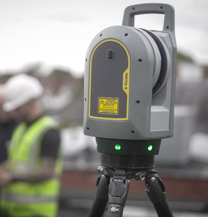 Grey surveying equipment with yellow accents and green LED lights mounted on black tripod, construction site background blurred.