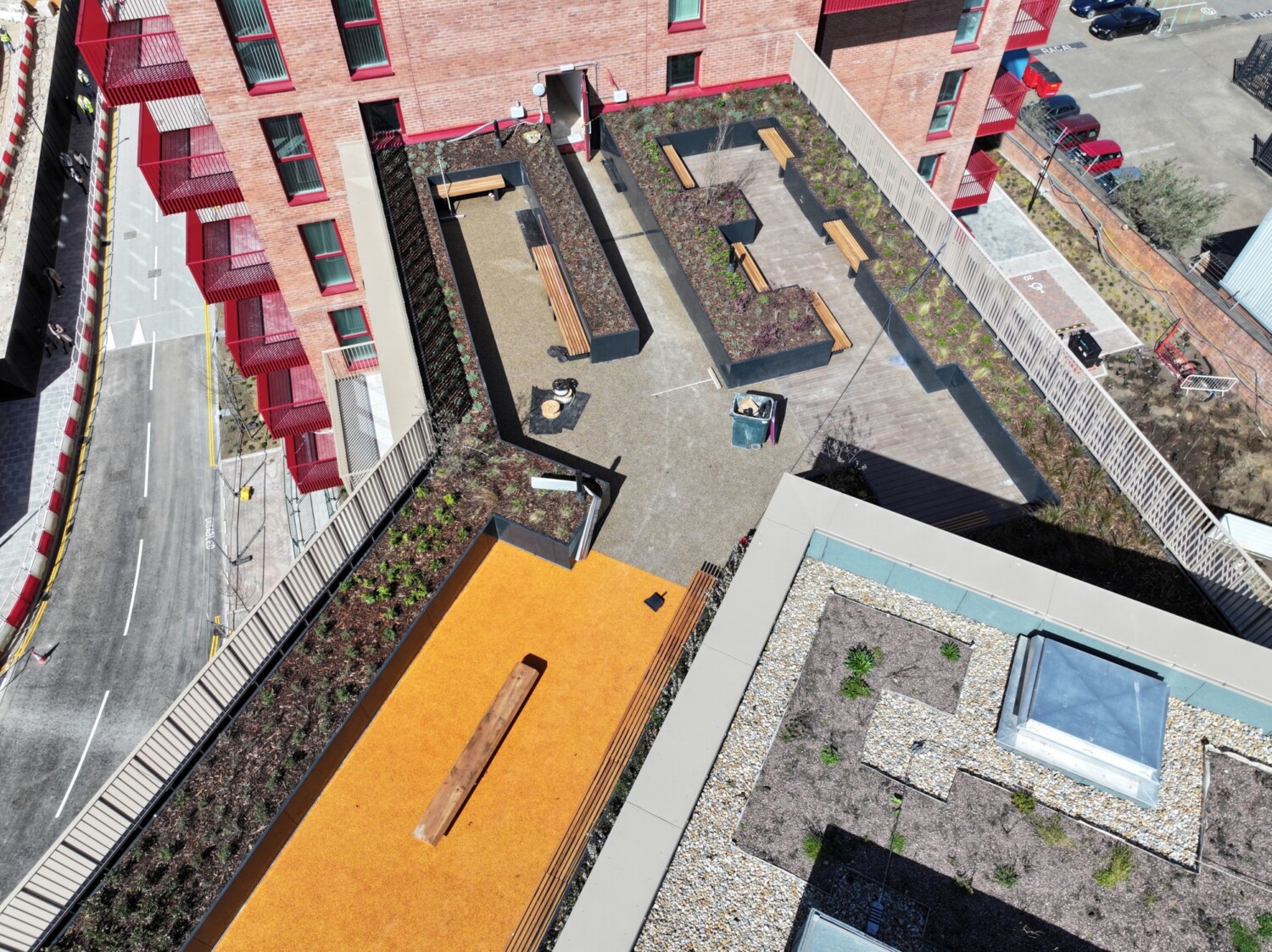 Aerial view of residential complex with orange rooftop, swimming pool, courtyard areas, and red-accented buildings along street.