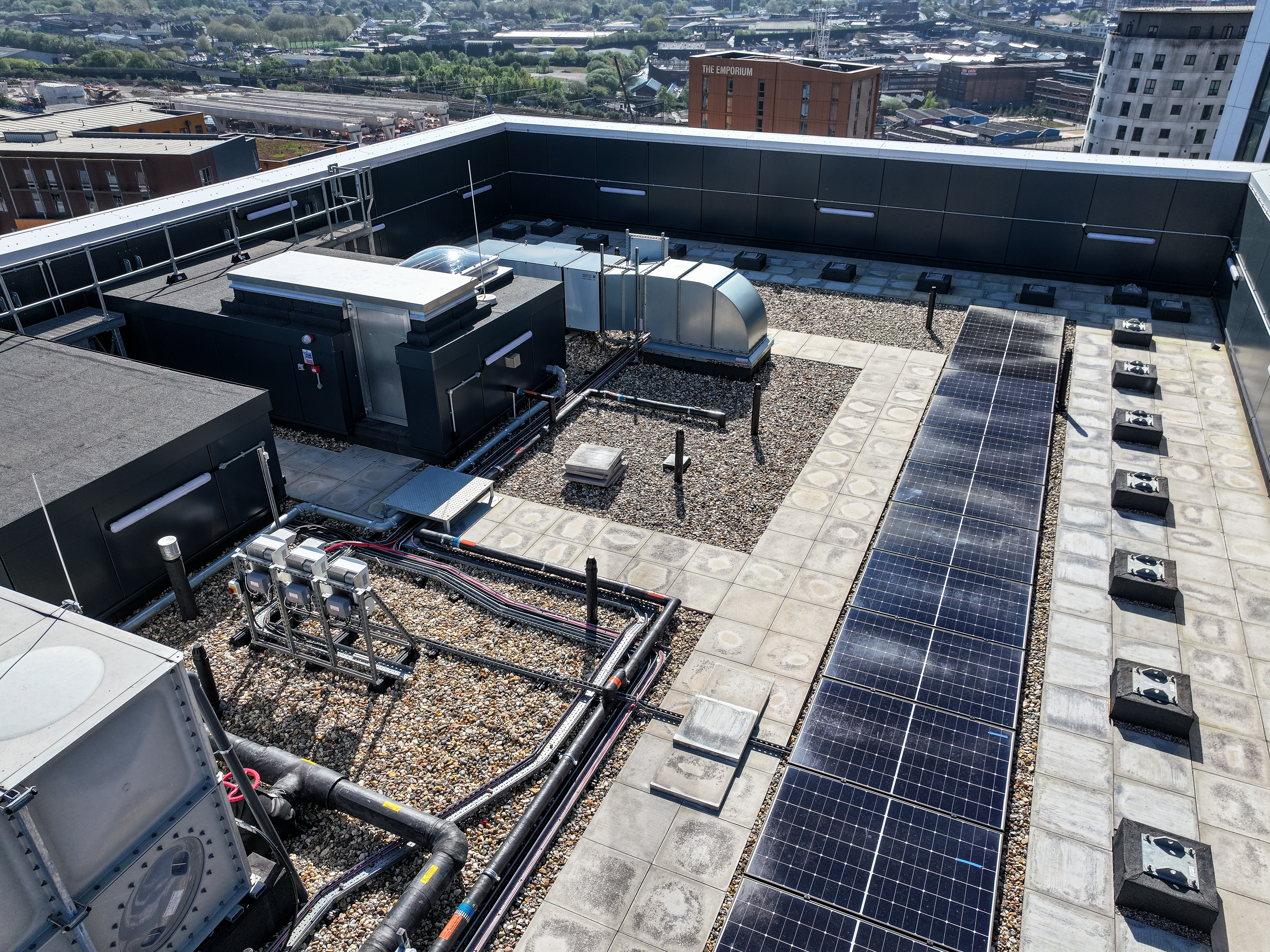 Aerial view of a flat rooftop with solar panels, HVAC units, pipework, and gravel surfacing. Urban skyline visible in the background.