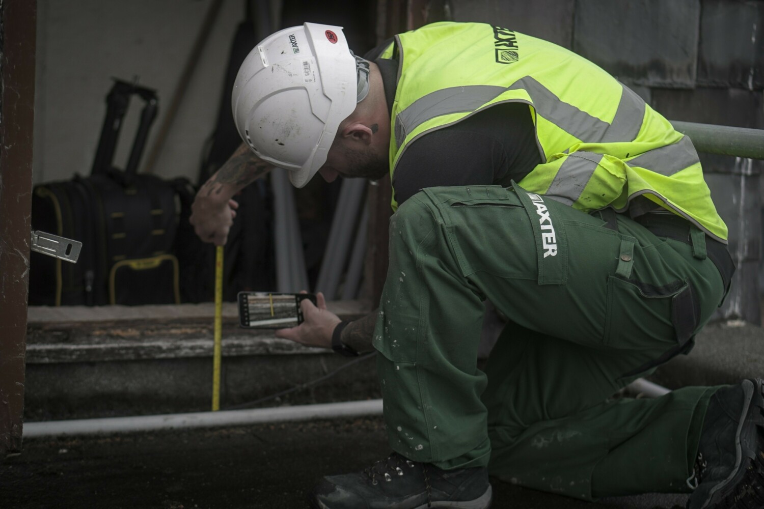 Worker in white hard hat and high-vis yellow jacket kneeling beside machinery, using measuring tool on metal surface in industrial setting.