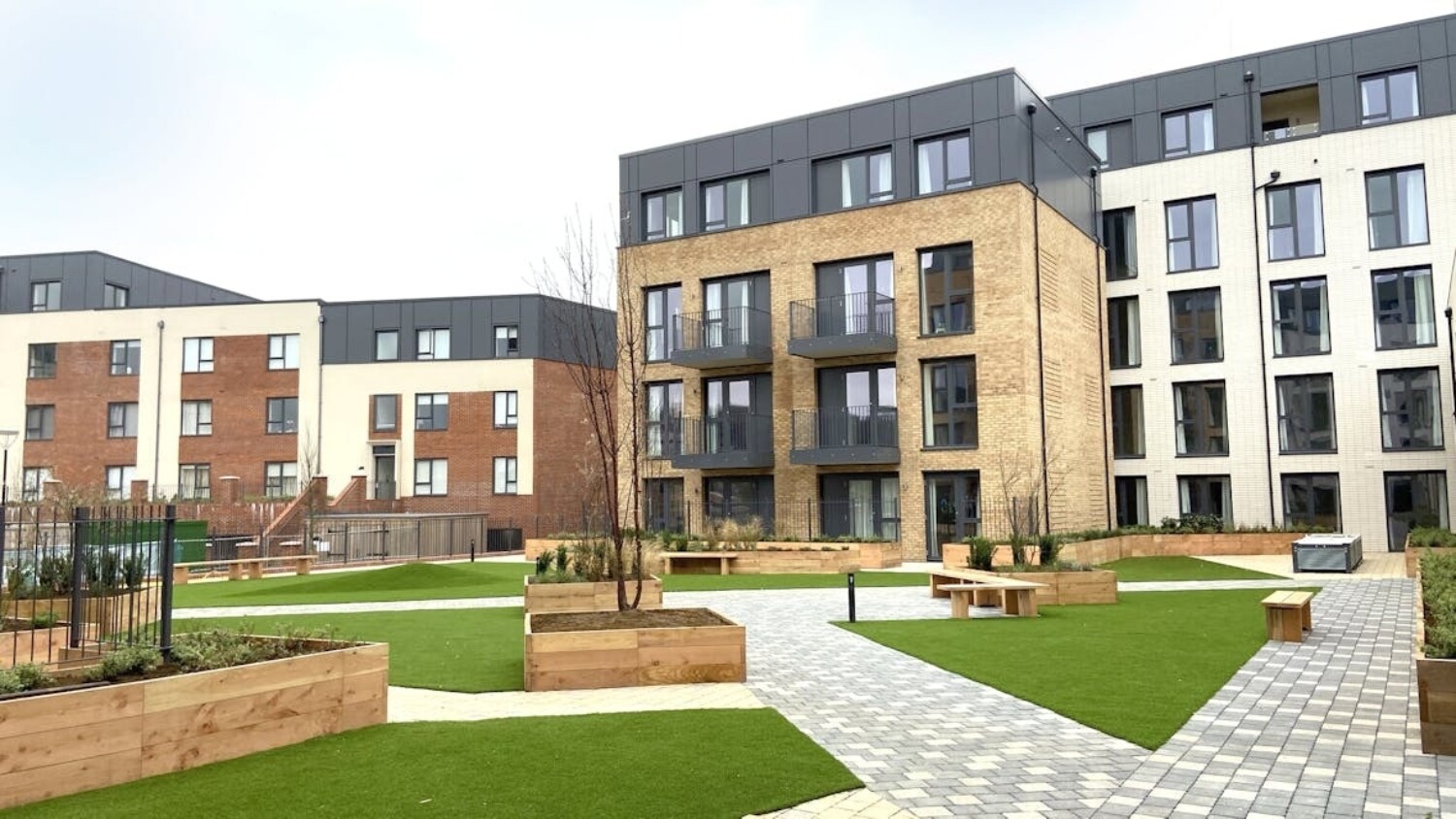 Modern residential development with multi-storey flats in brick, grey render and white facades surrounding landscaped courtyard with lawn areas and paved walkways.