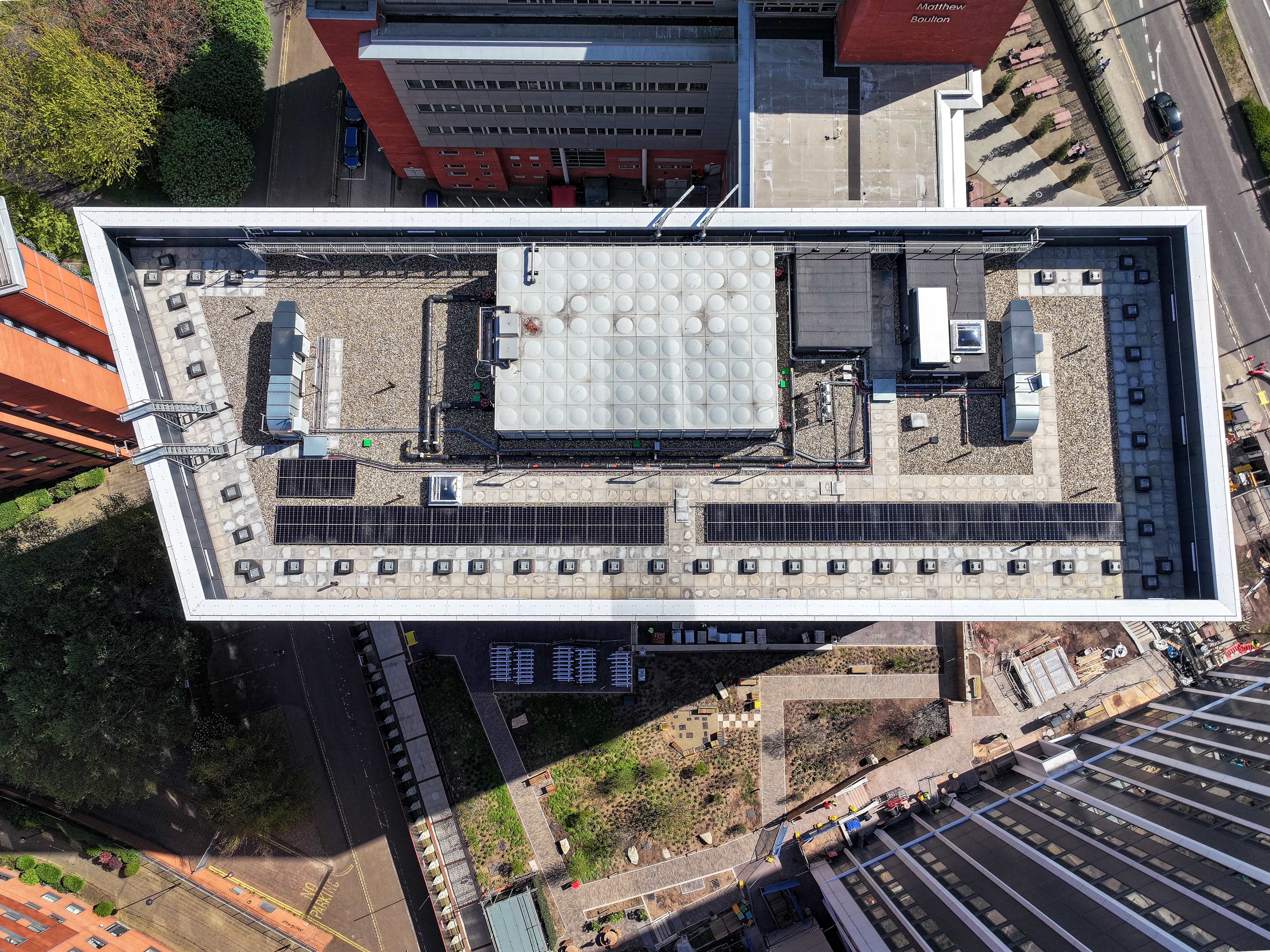 Aerial view of a flat commercial rooftop with solar panels, HVAC units, and a water tank, surrounded by urban streets and buildings.