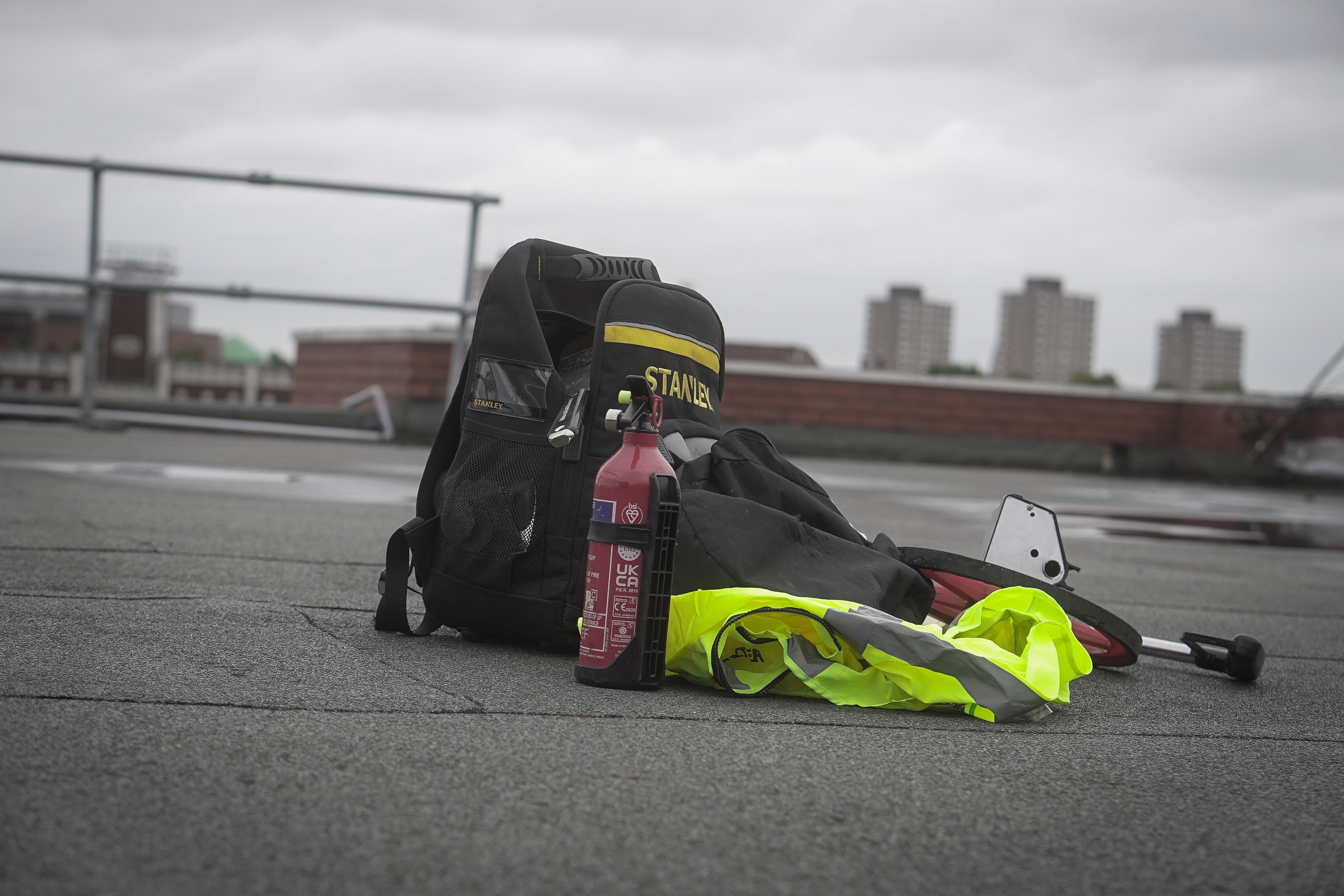 Black rucksack with high-visibility yellow jacket, pink water bottle, and other gear on concrete surface with city skyline background.
