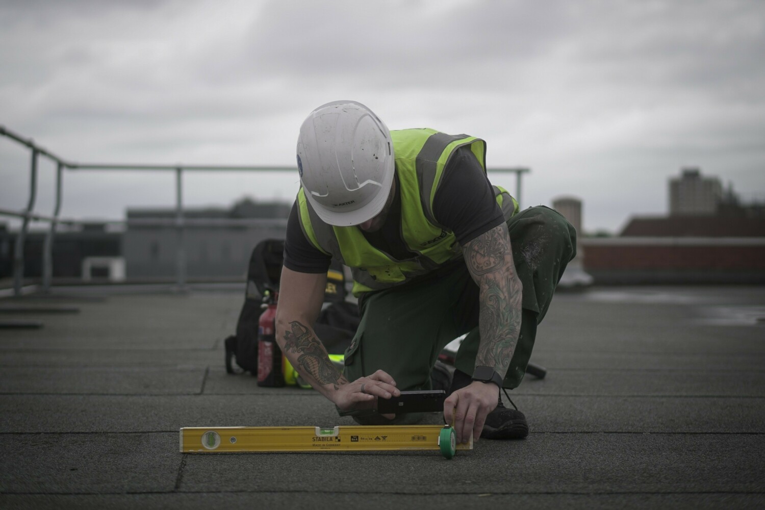 Construction worker in white hard hat and high-vis yellow jacket kneeling on rooftop, using spirit level on dark surface under cloudy sky.