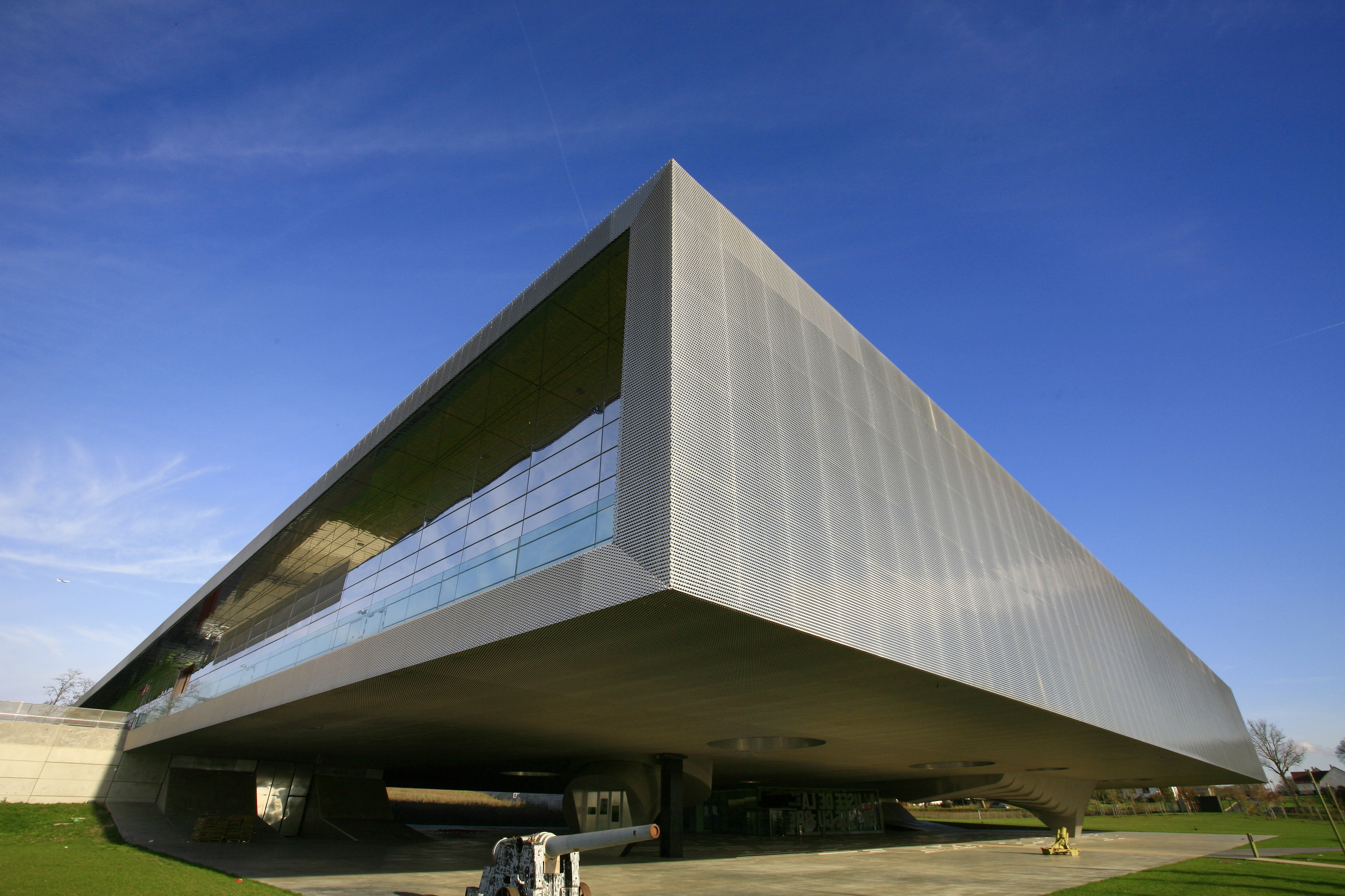 Modern angular building with white geometric facade and glass windows, featuring cantilevered upper section against blue sky.