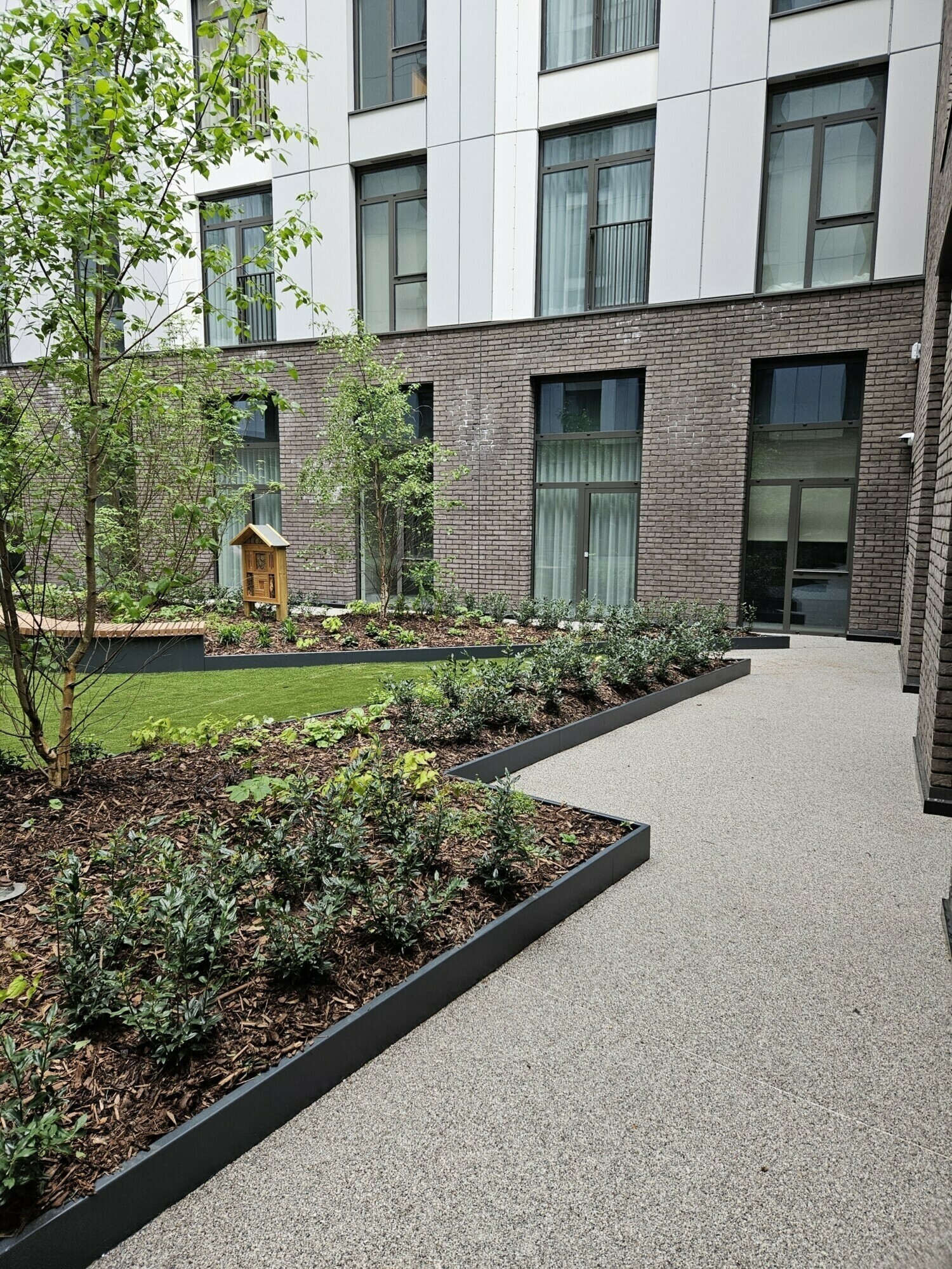 Modern residential building with white upper floors and brick ground level. Landscaped courtyard with raised planters, lawn, and trees.