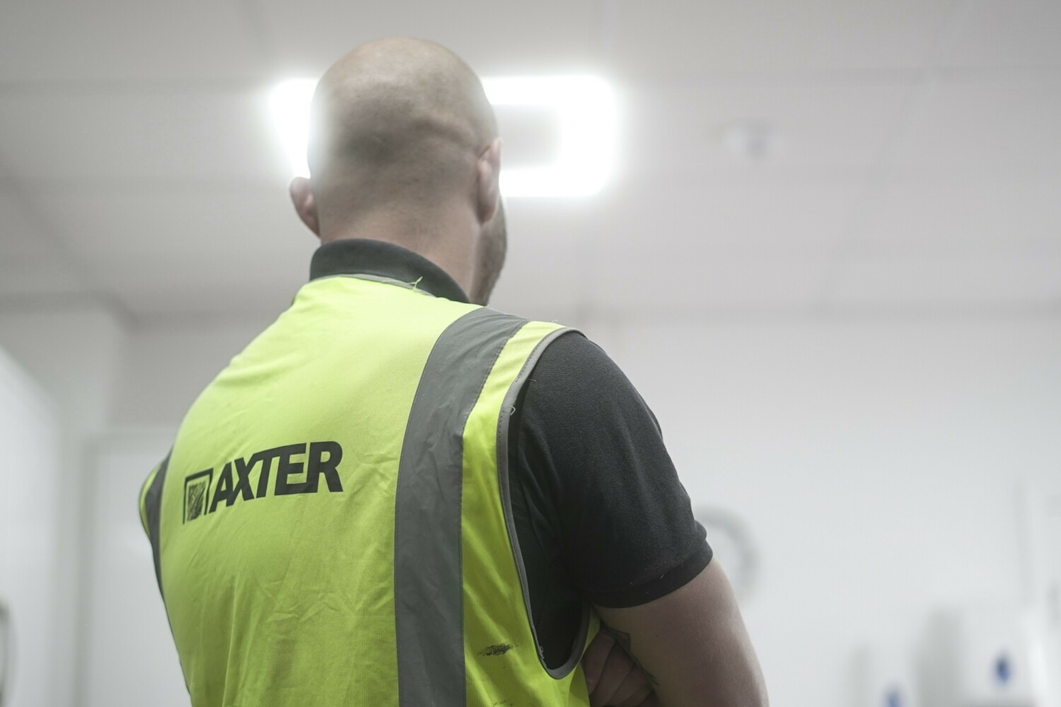Man in yellow high-visibility vest with "BAXTER" text and reflective stripes, viewed from behind in industrial setting.