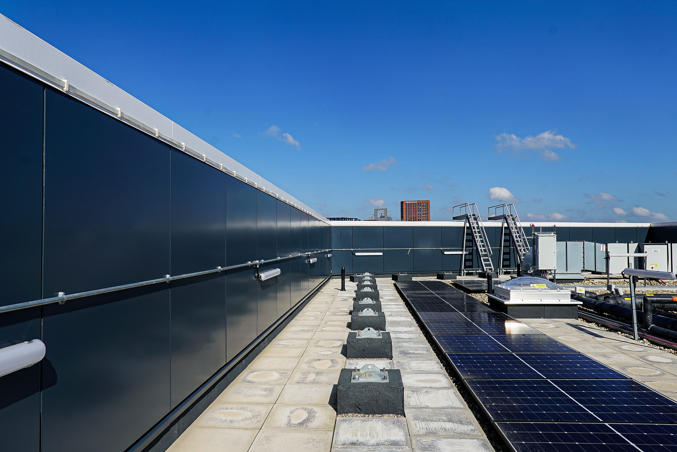 Flat rooftop with solar panels, dark cladding wall, concrete pavers, rooftop equipment, and a clear blue sky with a city building in the background.