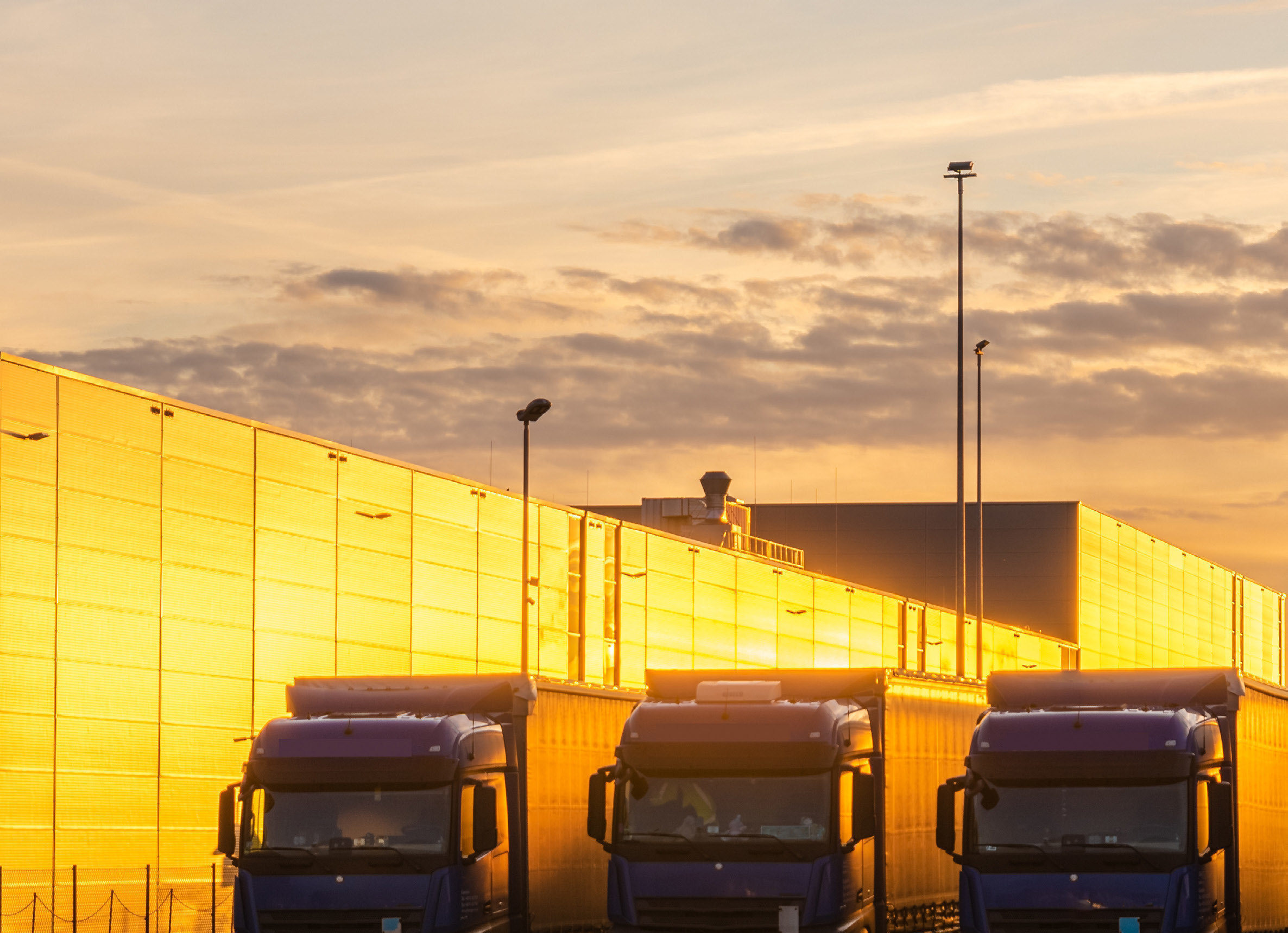 Three lorries parked at loading bays of bright yellow warehouse building under orange and purple sunset sky with lamp posts.