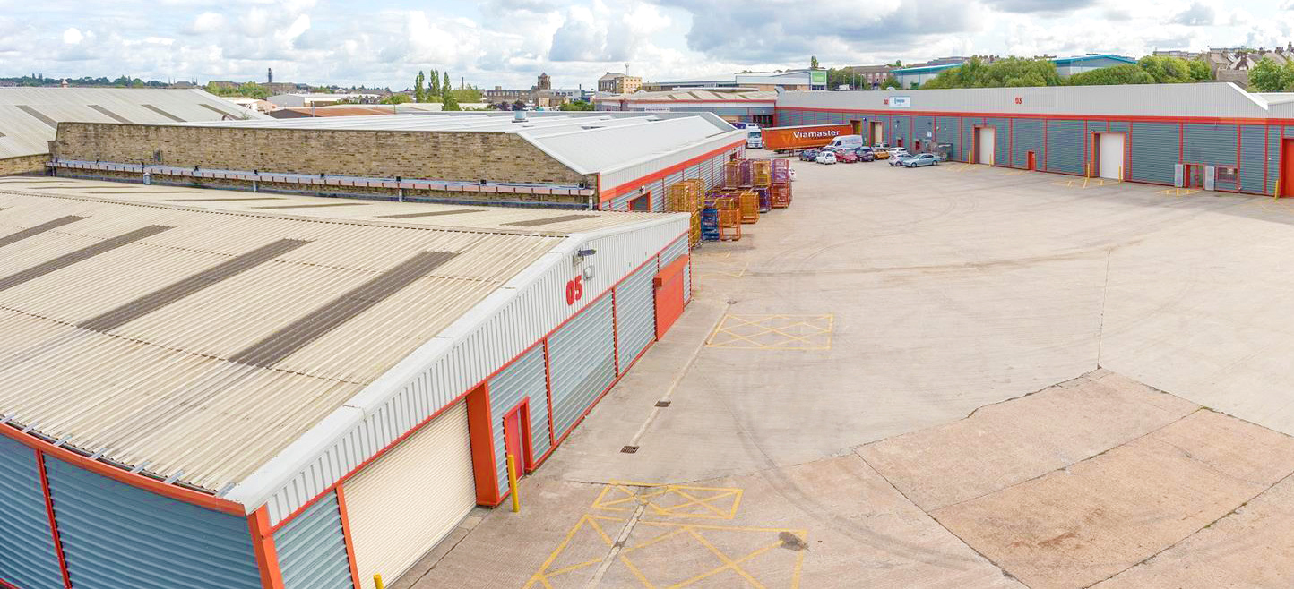 Industrial estate with corrugated metal warehouses, red trim, concrete yards, and cloudy sky overhead.