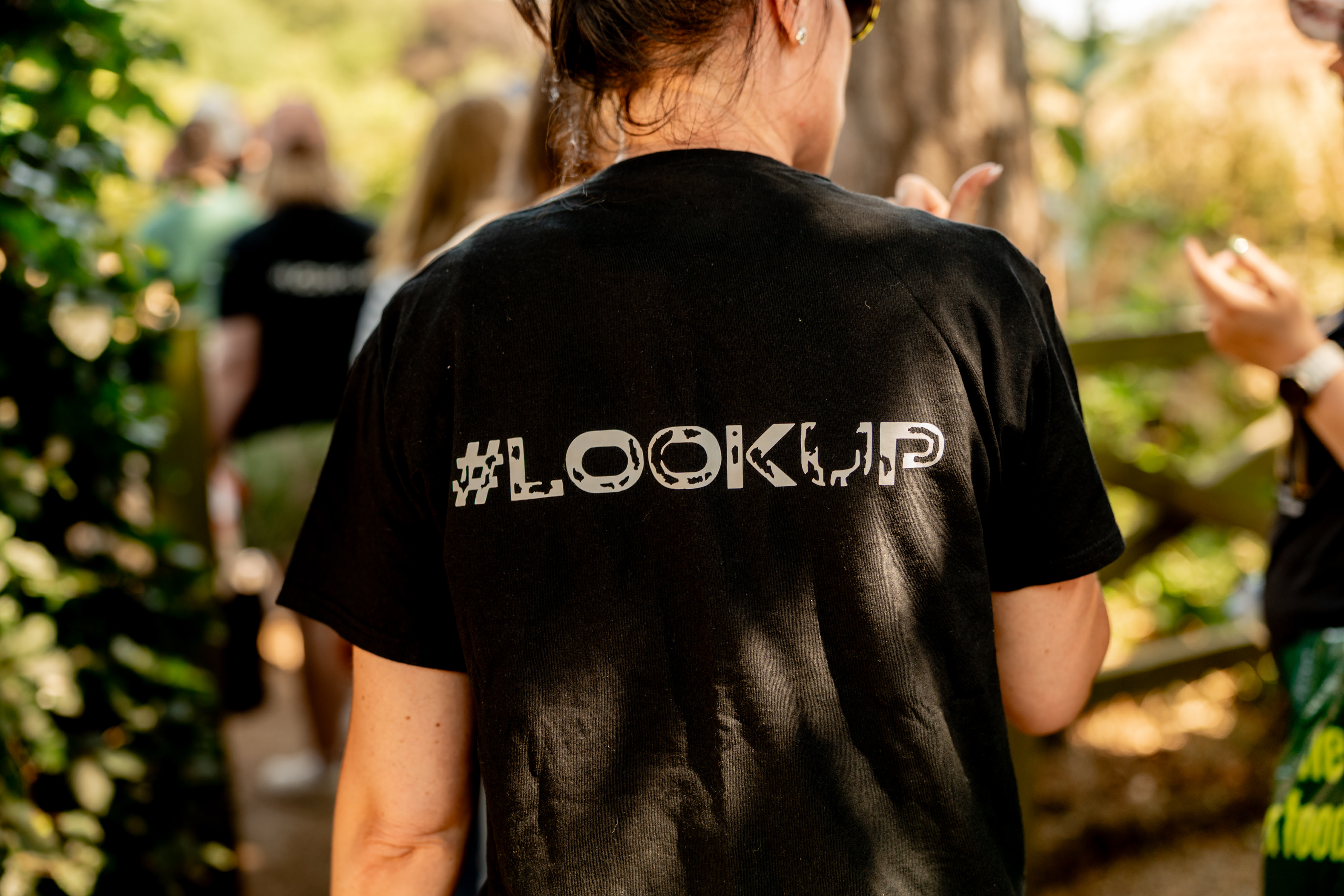 Person seen from behind wearing a black t-shirt with '#LOOKUP' printed on the back, standing outdoors among trees and other people.