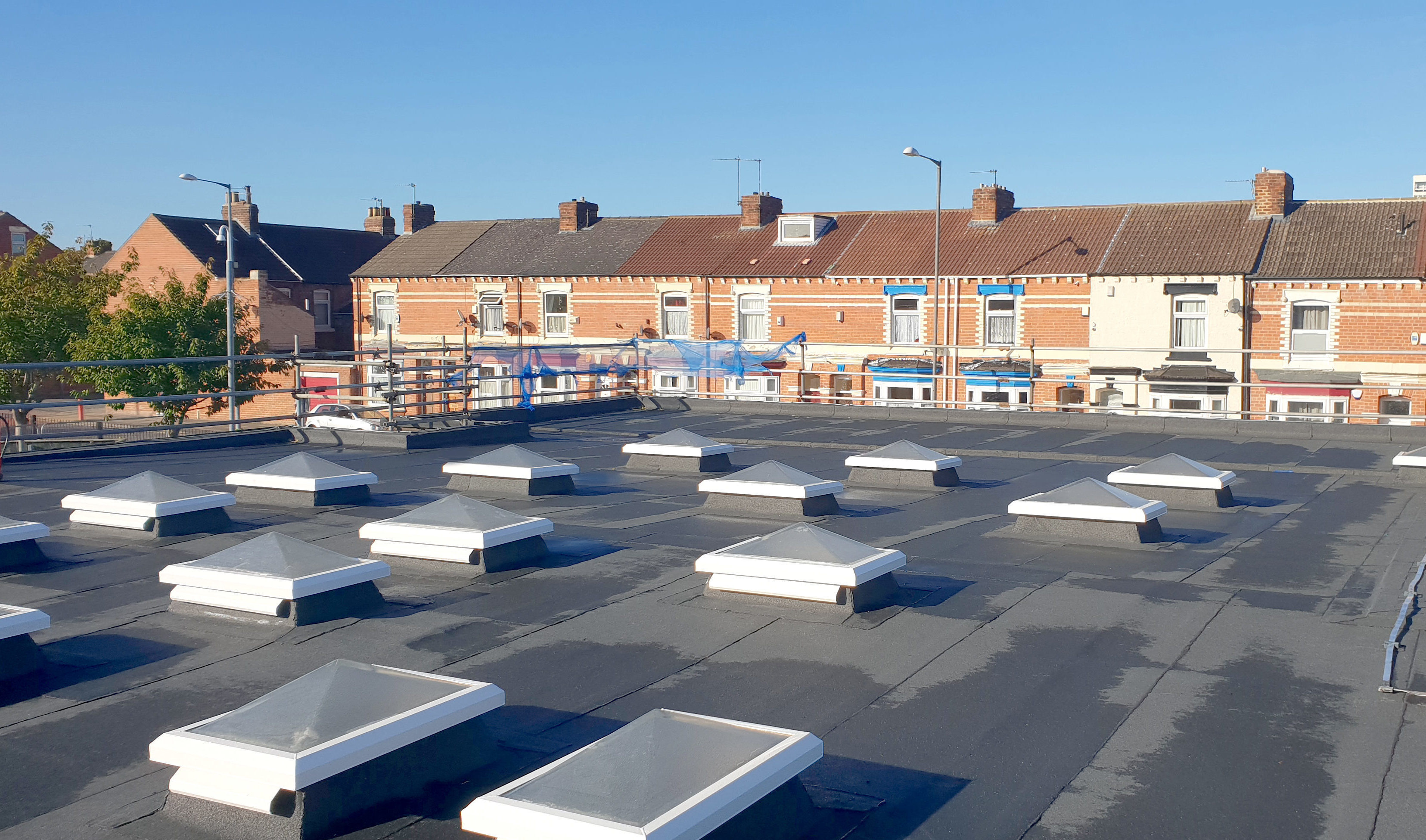 Flat roof with white rectangular ventilation units arranged in rows, terraced houses with red brick and coloured doors in background.