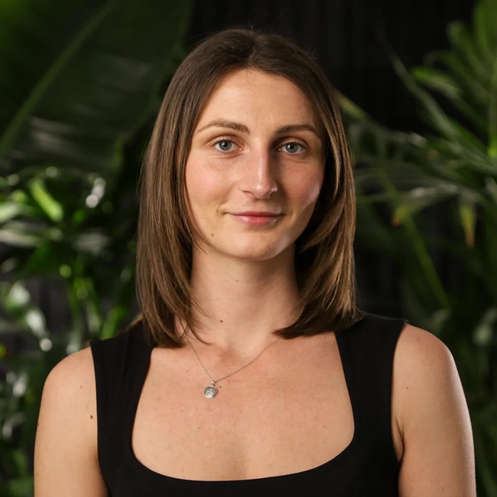 Woman with shoulder-length brown hair wearing black sleeveless top and small necklace, smiling at camera with green foliage background.