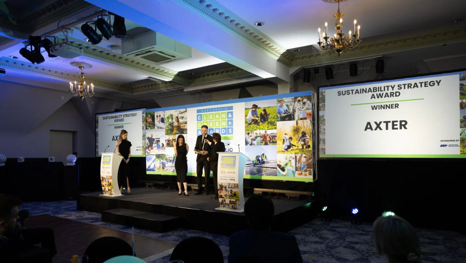 Conference hall with three people on stage presenting sustainability awards. Large projection screen displays "AXTER" branding and presentation materials. Ornate ceiling with chandeliers, audience seated in foreground.