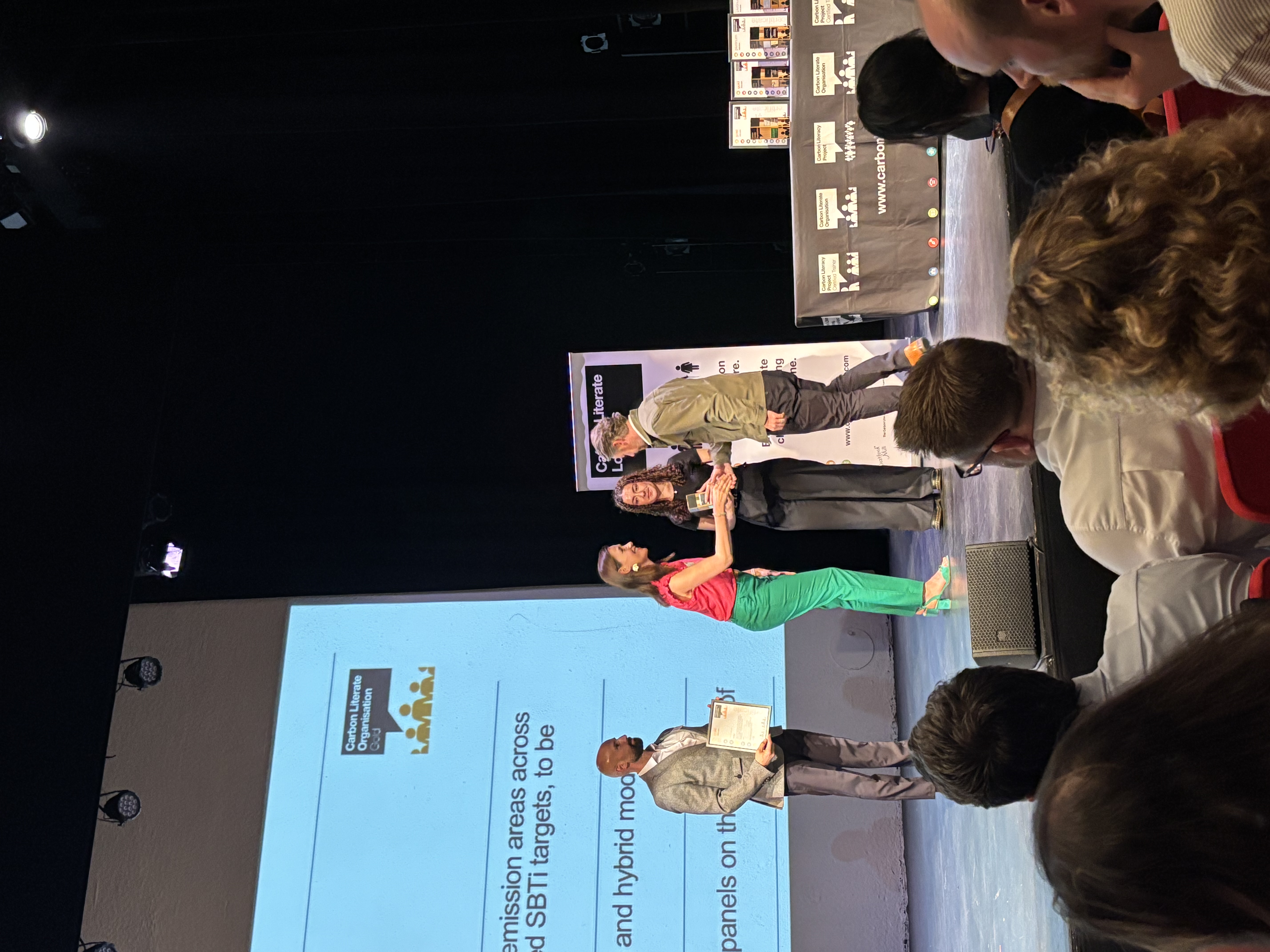 Woman in green dress and red cardigan presenting at podium with blue projection screen, audience members visible in foreground.