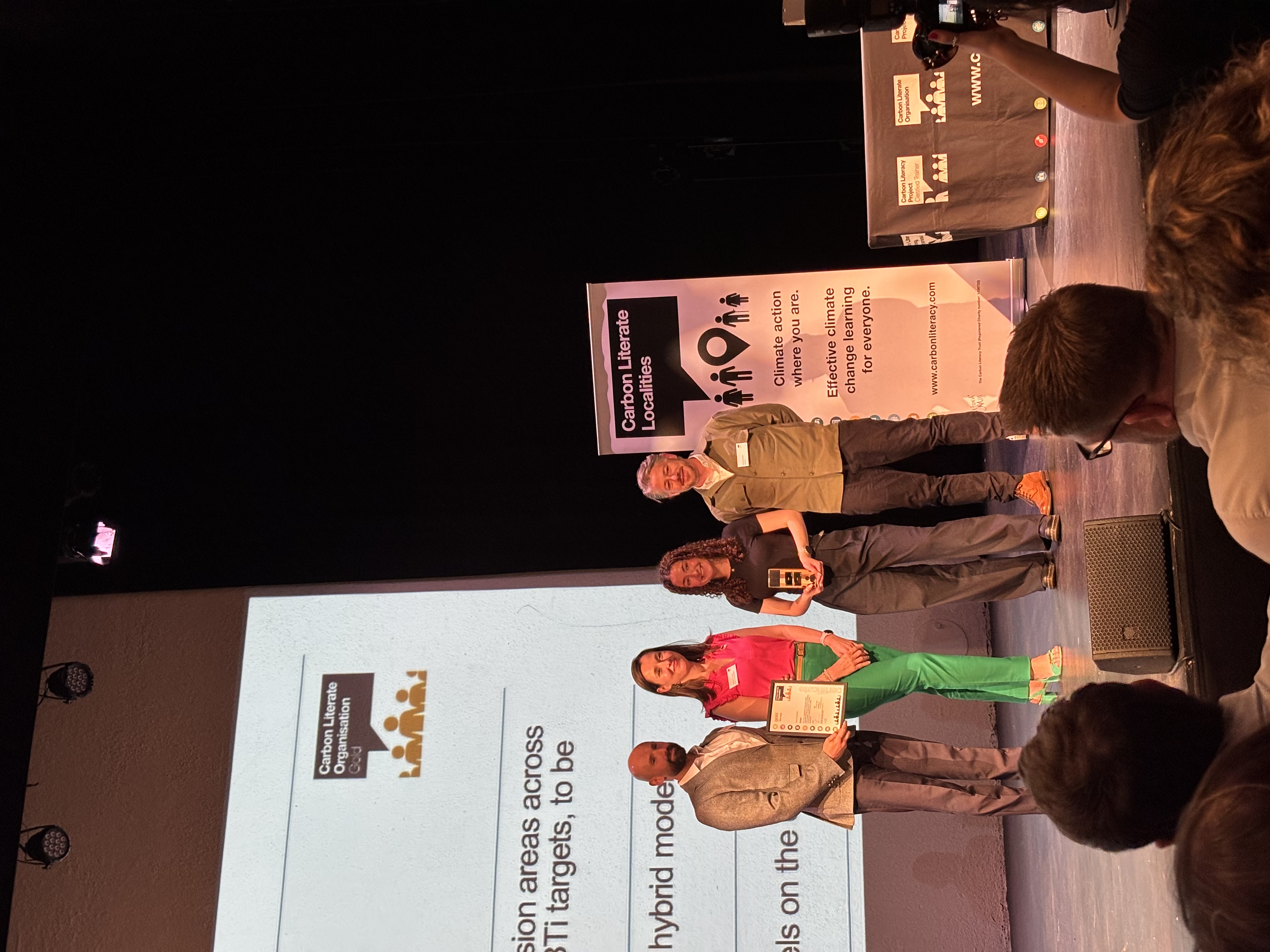 Four people standing on stage beside large presentation screen displaying text about hybrid models at tech conference event.