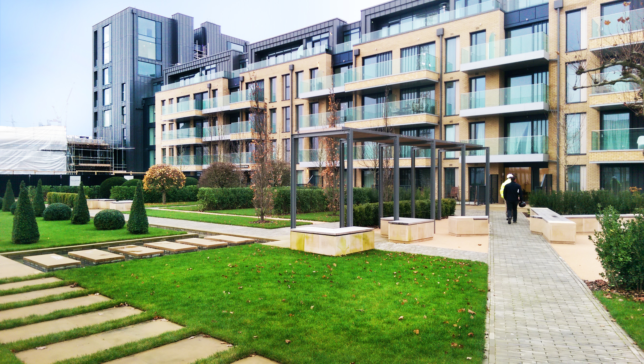 Modern residential complex with brown brick and glass facades, landscaped courtyard with green lawn, paved pathways, and geometric planters.