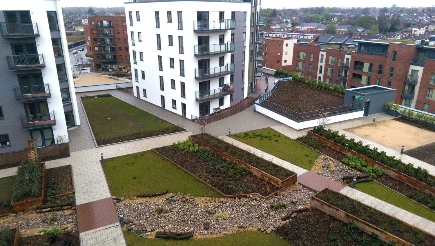 Modern residential development with white and brick apartment blocks surrounding landscaped courtyard featuring green lawns, planted beds, and paved walkways.