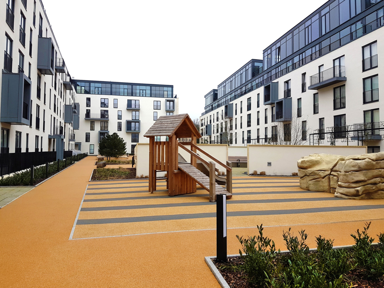 Modern residential development with white and grey apartment blocks surrounding children's playground with wooden climbing frame and orange safety surfacing.