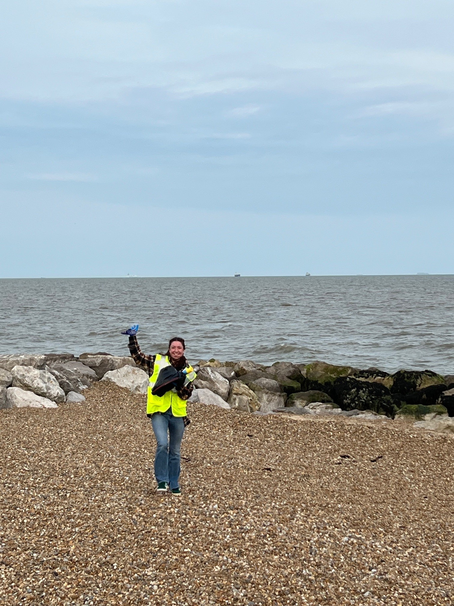 Person in bright yellow high-vis jacket standing on pebble beach, giving thumbs up. Stone breakwater behind, grey sea and cloudy sky.