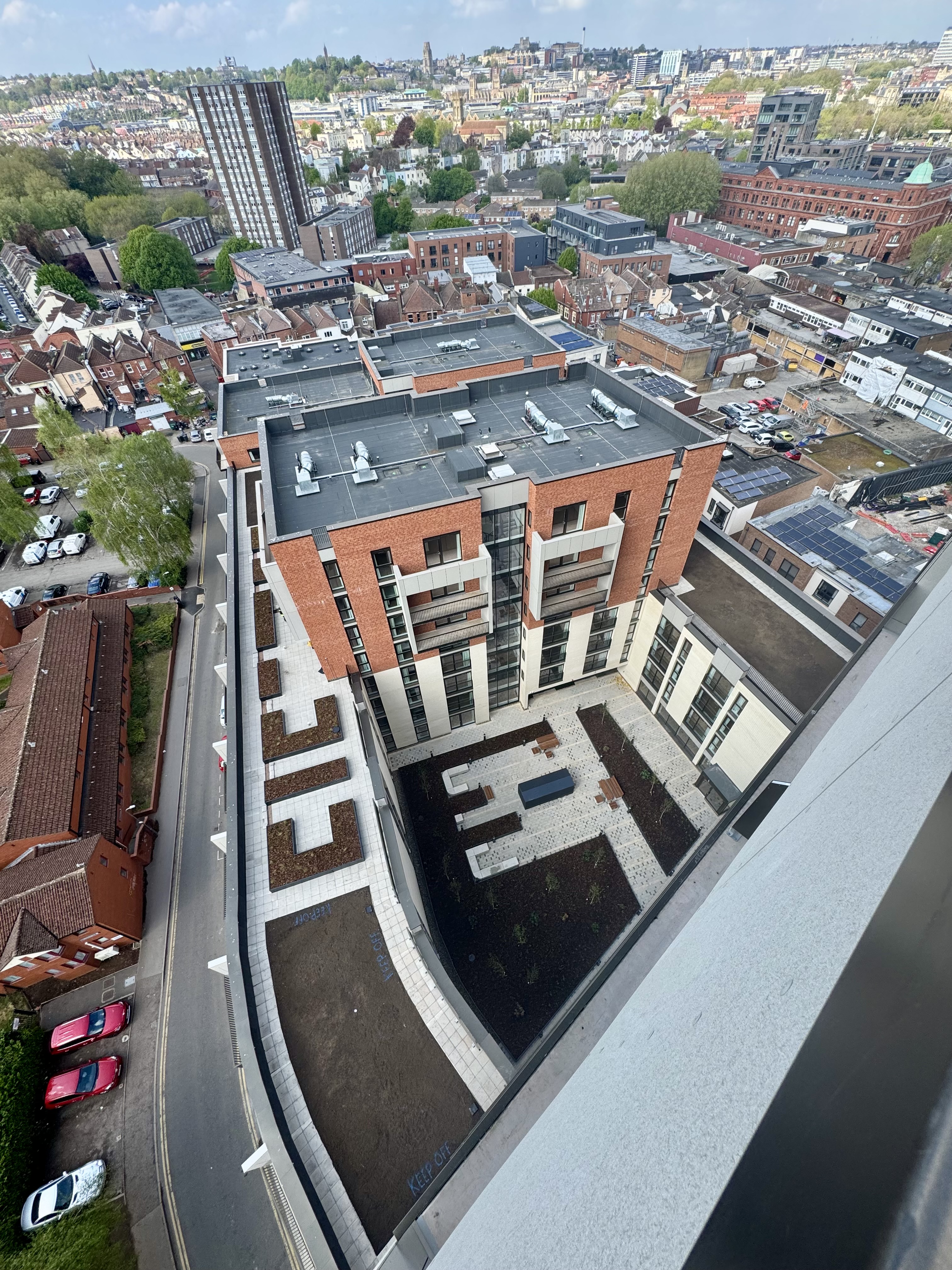 Aerial view of modern multi-storey residential building with red brick and white facades, surrounded by urban streets and housing.