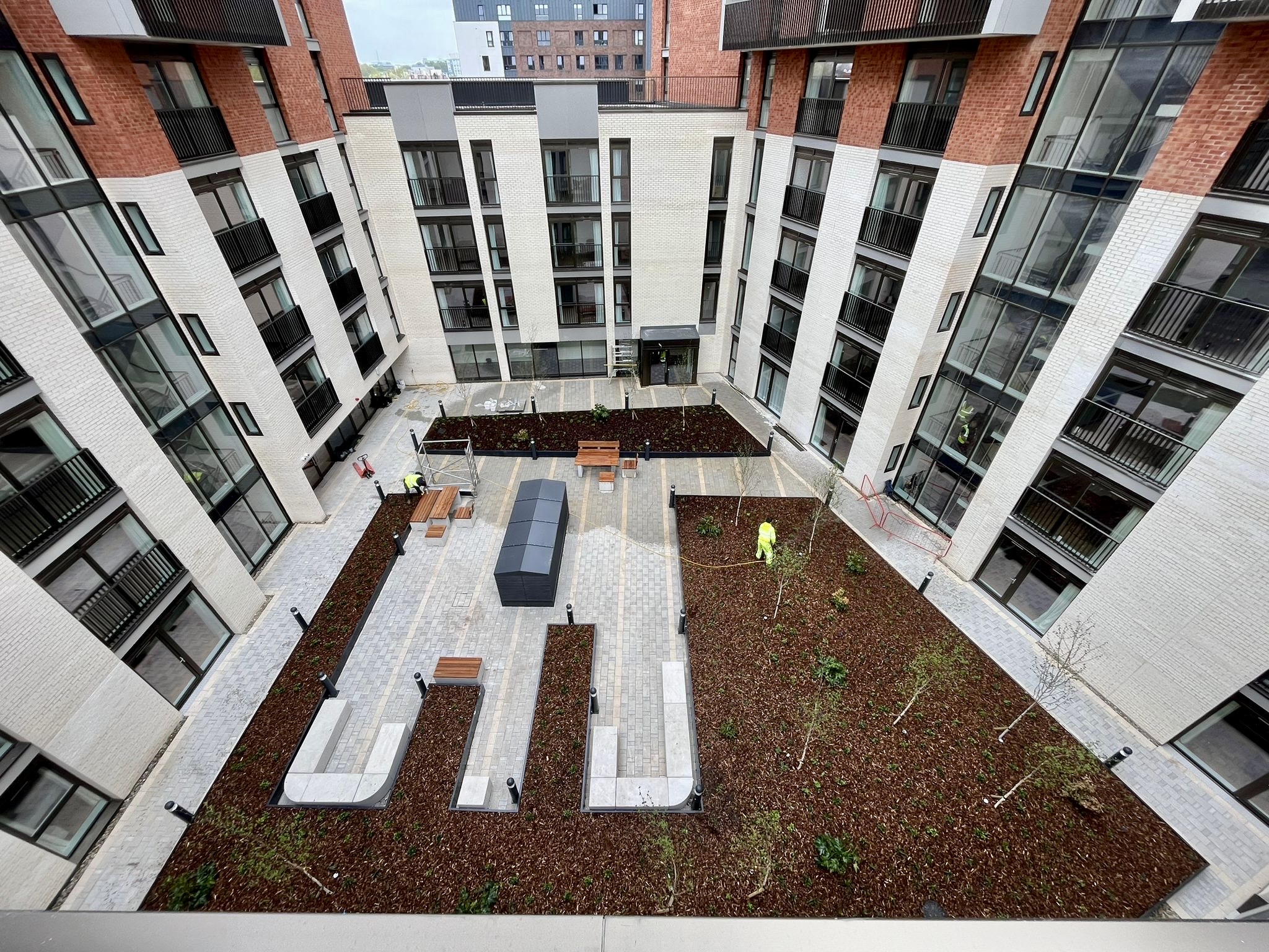 Aerial view of rectangular courtyard surrounded by modern flats with red brick and white facades, featuring landscaped garden beds and seating.