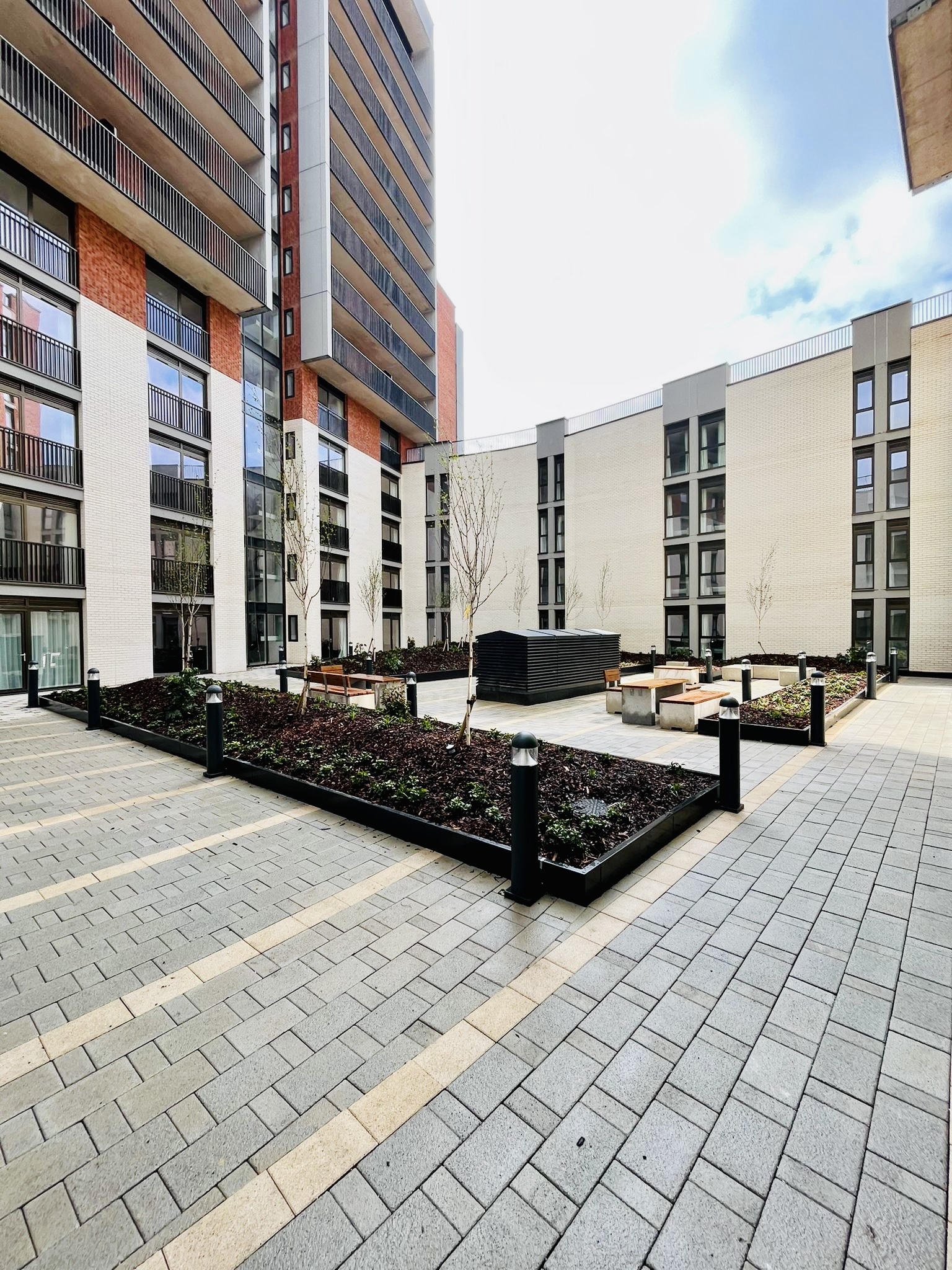 Modern residential courtyard with grey block paving, raised planters with dark soil, wooden benches, and surrounding multi-storey flats with balconies under cloudy sky.