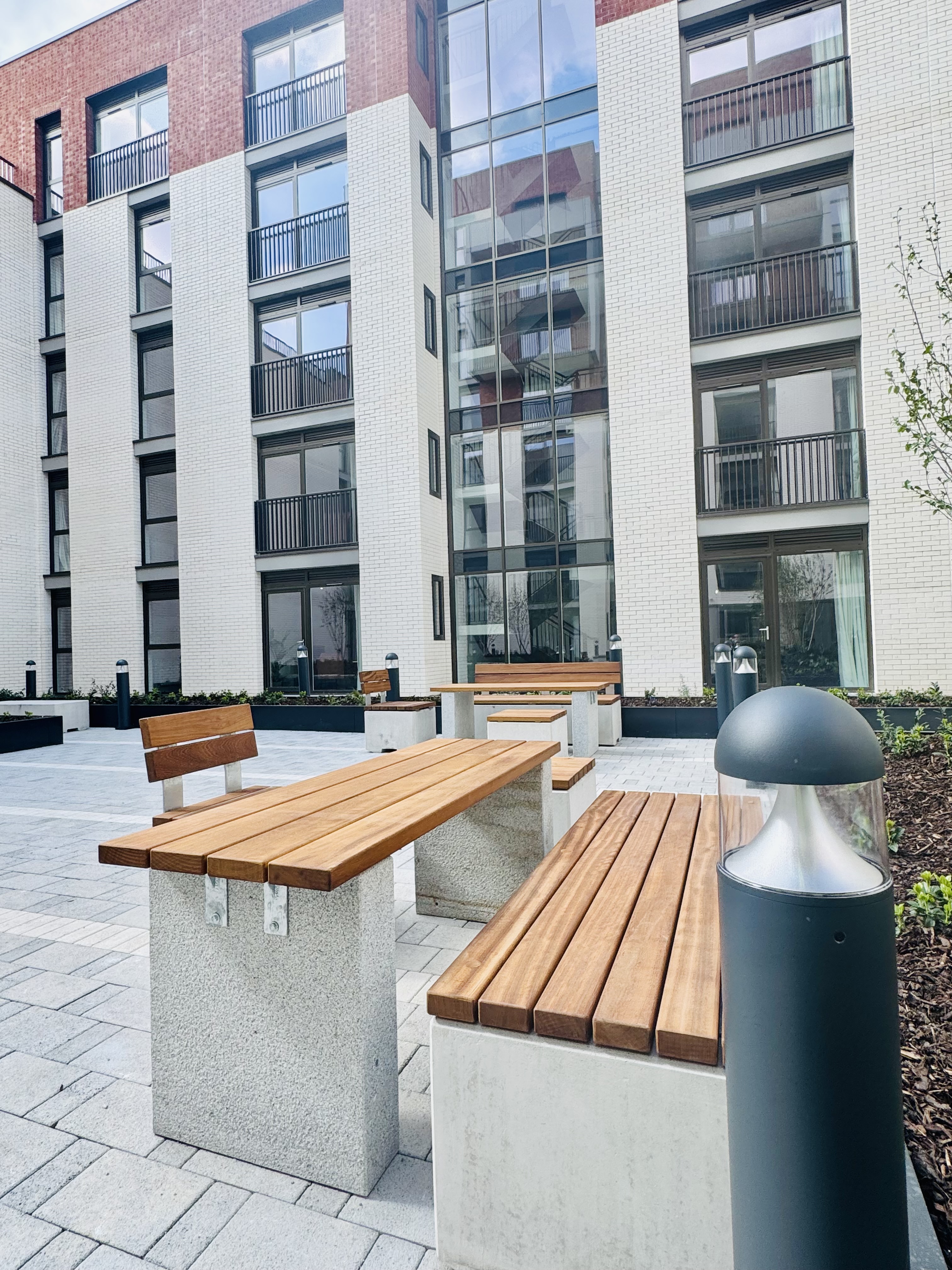 Modern residential courtyard with white and red brick buildings, wooden bench seating on concrete bases, and cylindrical bollard lighting.