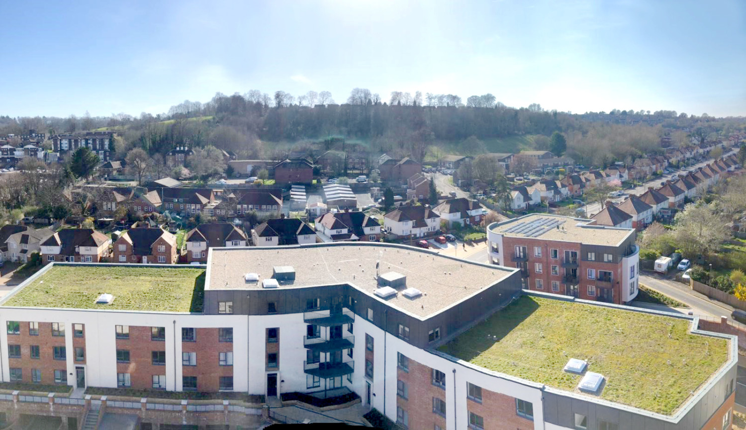 Aerial view of modern residential development with flat green roofs in foreground, traditional houses on hillside beyond under blue sky.