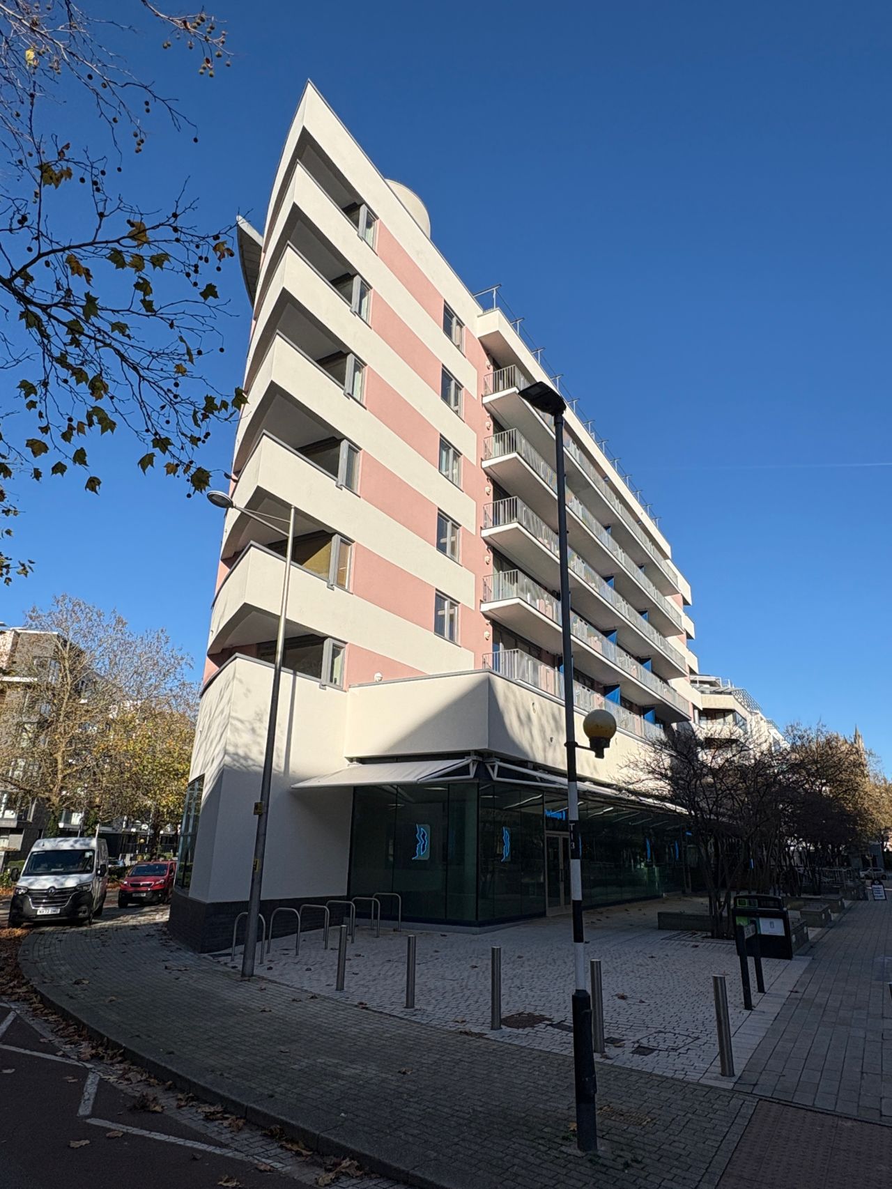 Modern apartment building with white and pink striped facade, multiple balconies, ground floor shops, bare trees and blue sky.