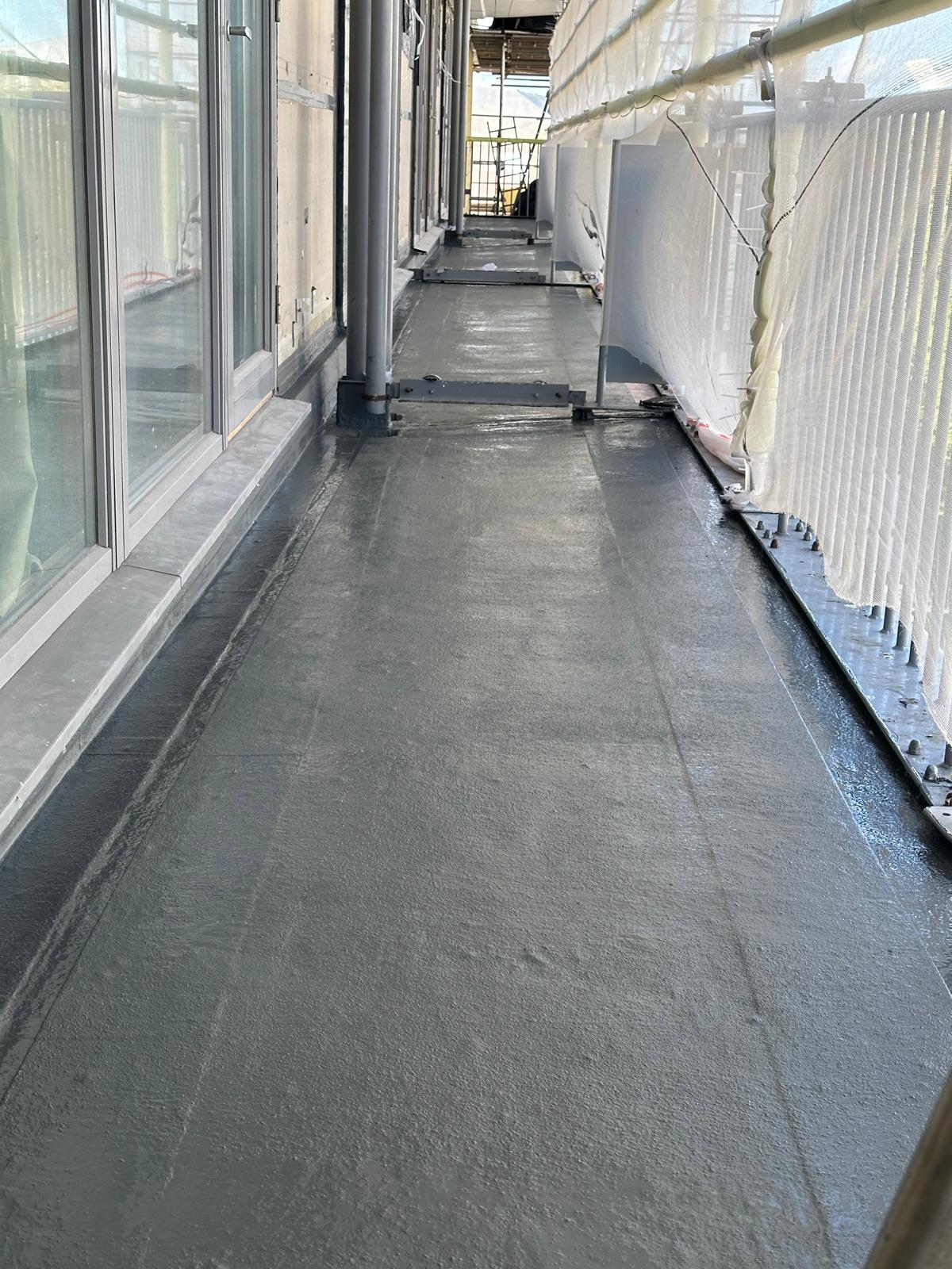 Narrow balcony walkway with grey flooring, glass windows on left, white corrugated metal sheeting on right, construction materials visible.