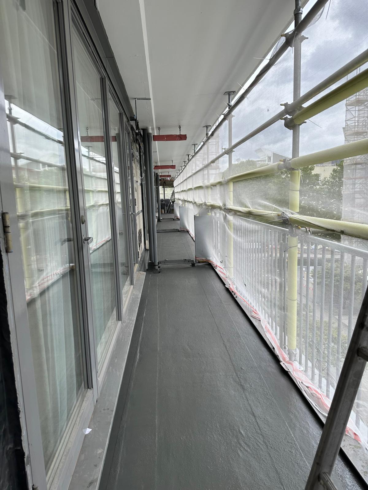 Narrow outdoor walkway with glass windows on left, white metal railings on right, grey flooring, and scaffolding visible outside.