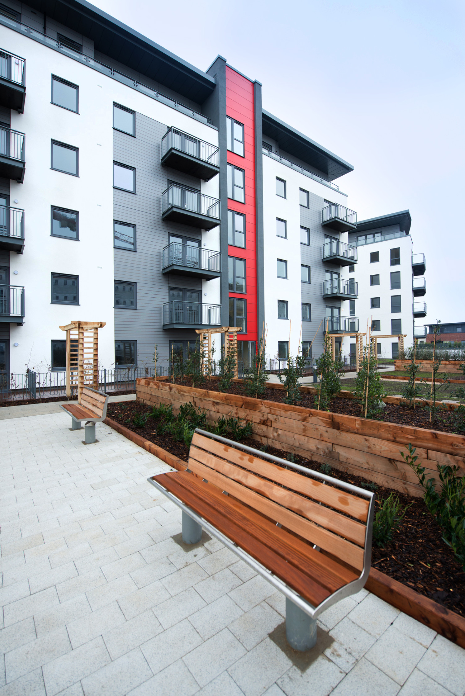 Modern apartment buildings with white walls, grey cladding, and red accent panel. Wooden bench on paved courtyard with raised planters.