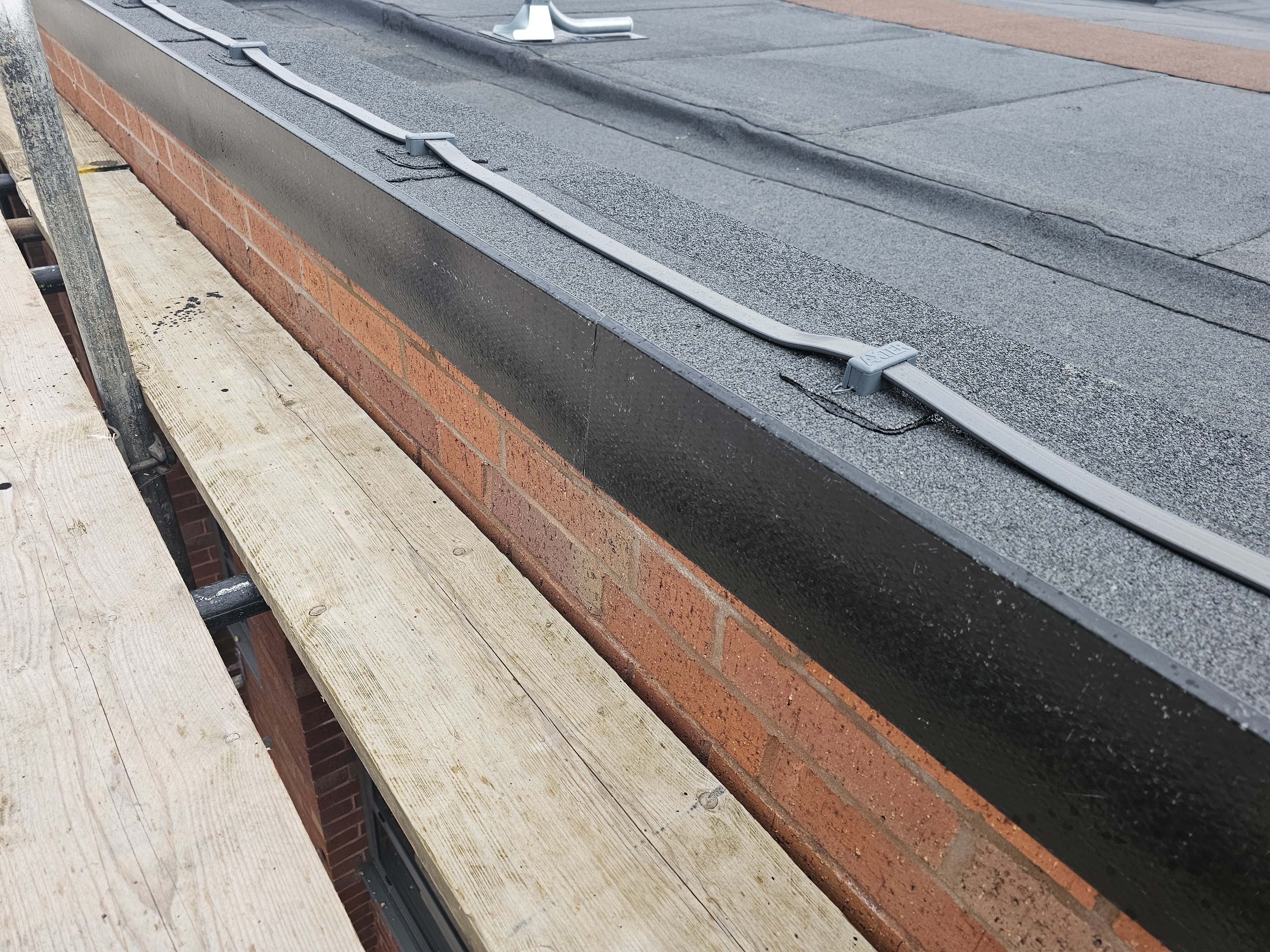 Grey flat roof with metal clips and guttering alongside red brick wall and wooden planks below.