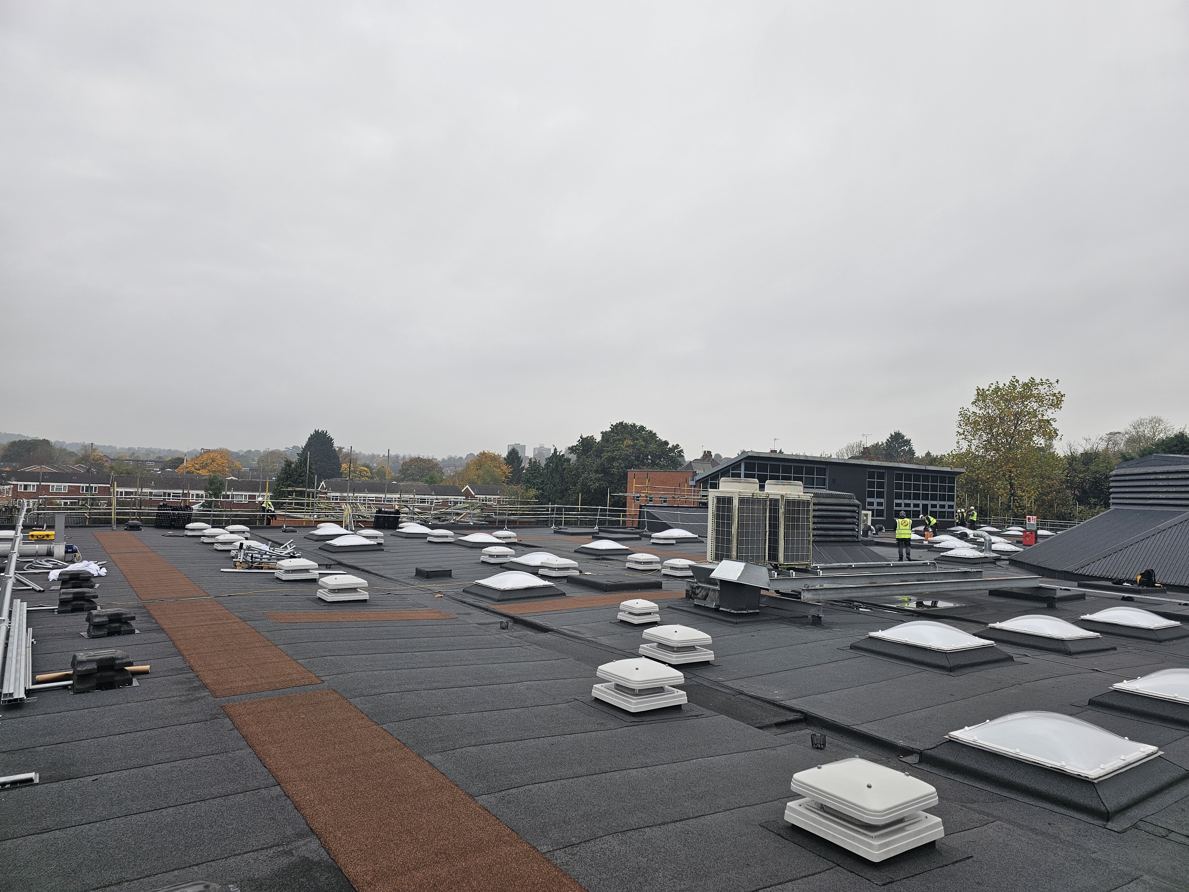 Flat roof with white ventilation units, HVAC equipment, and orange safety walkway. Trees and buildings visible in distance under grey sky.