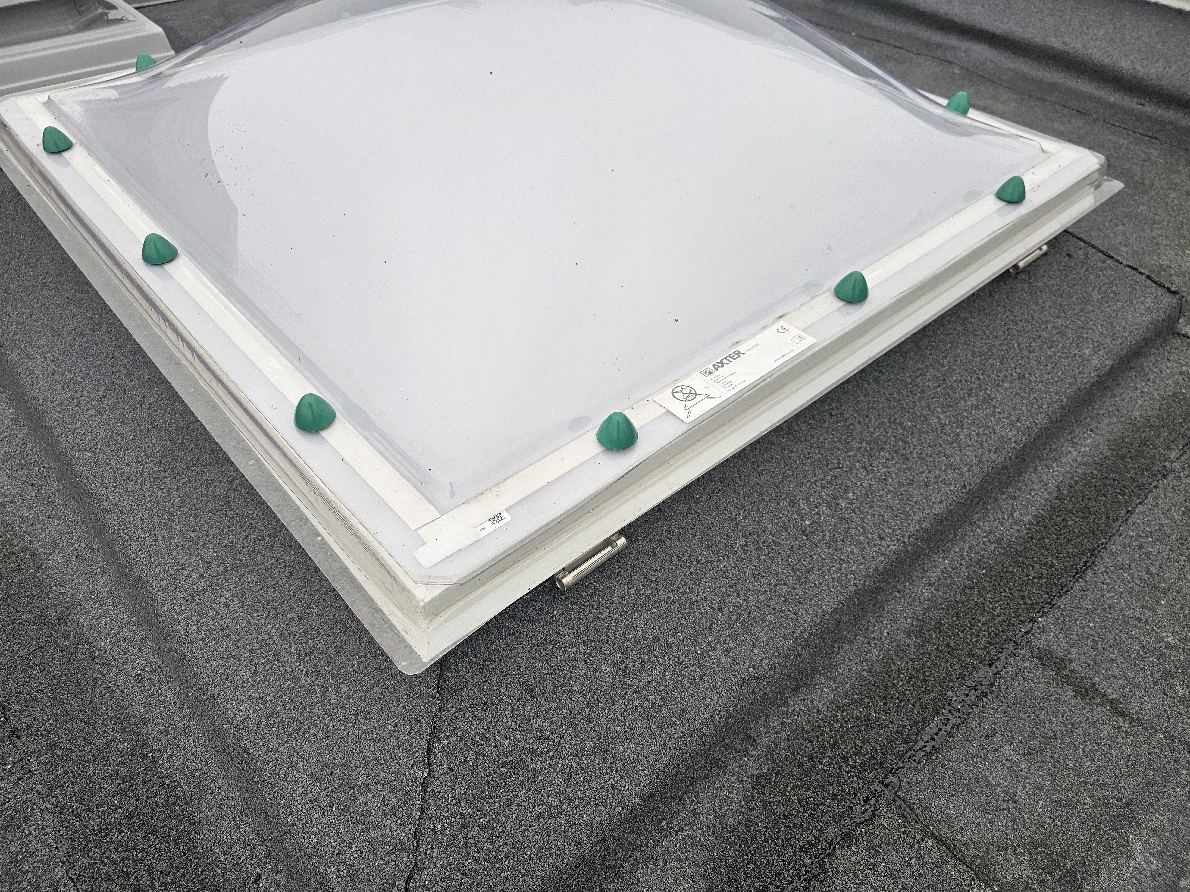 White rectangular skylight with green suction cup lifting devices positioned around edges, placed on grey rooftop surface.