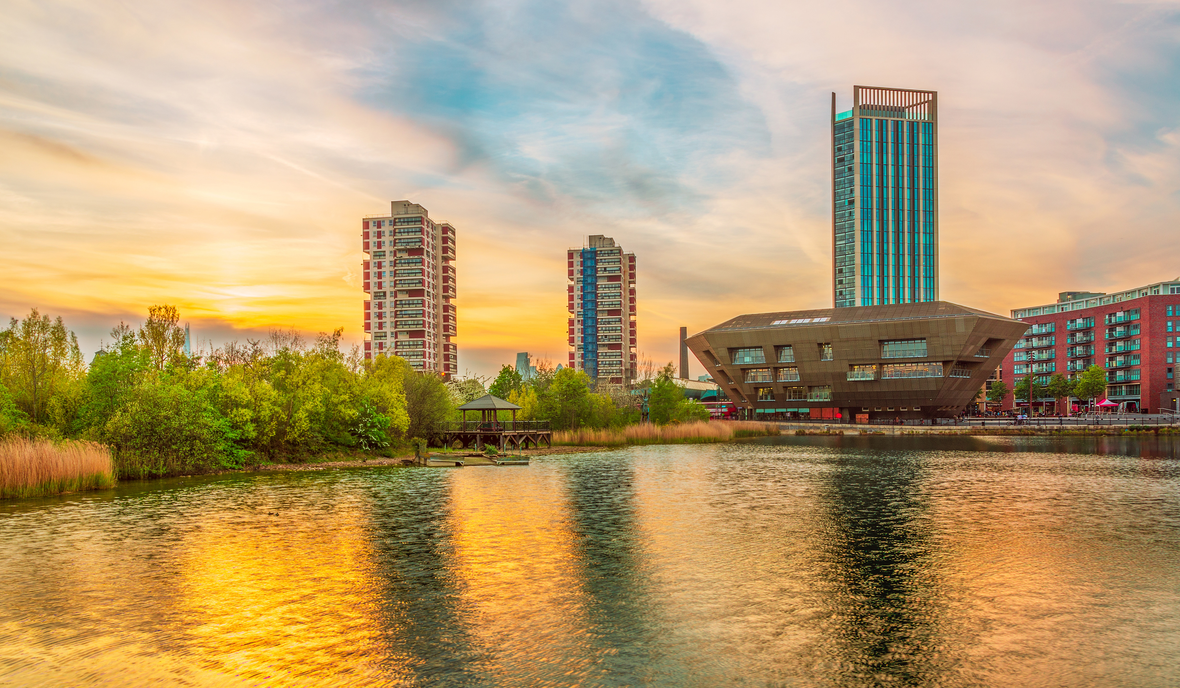 Urban waterfront with tall buildings reflected in river, green vegetation on left bank, orange and blue sky with clouds above.